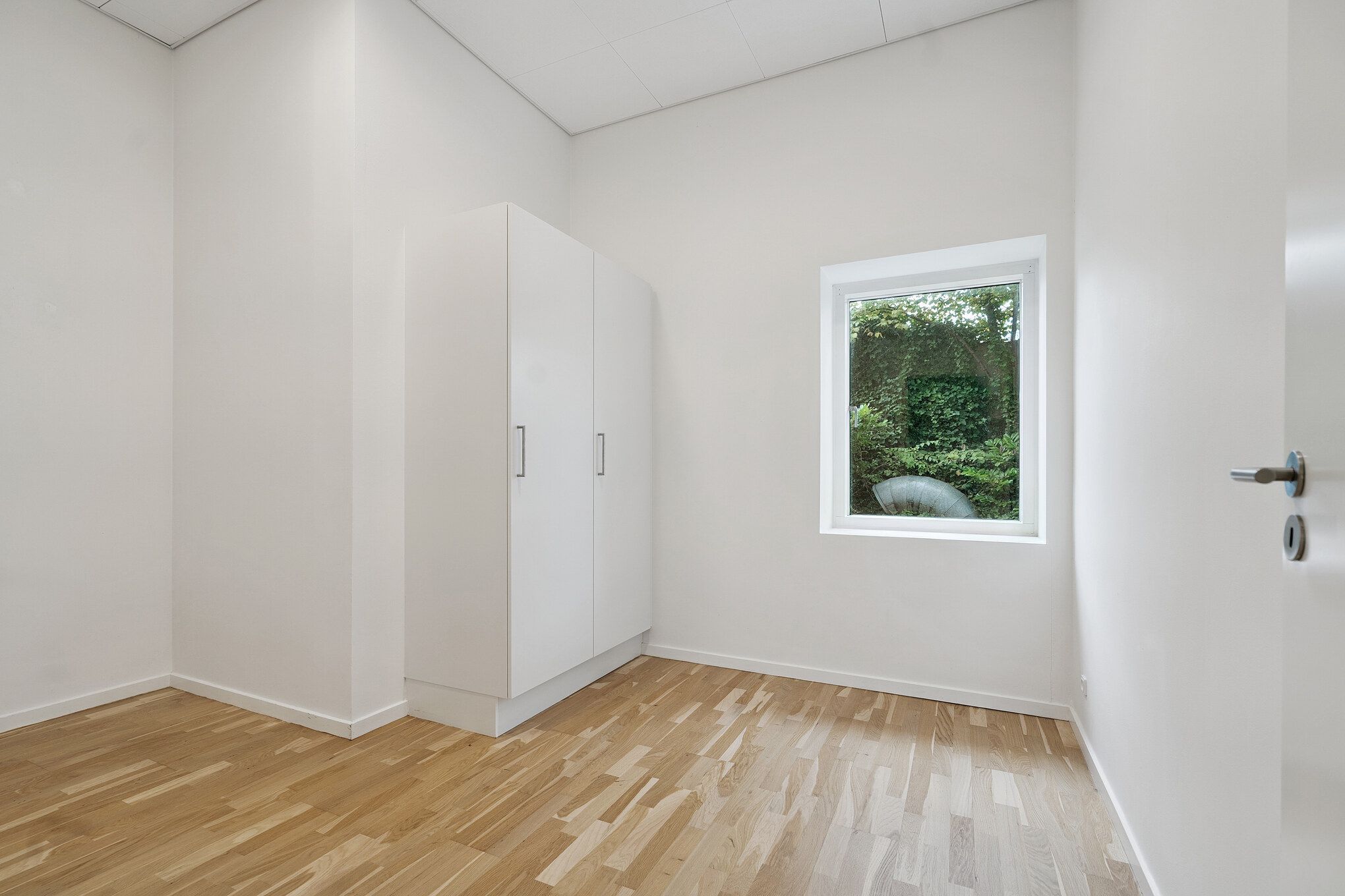 Funktionelt kontorrum med hvide vægge og trægulv. Der er et vindue med udsigt til grønne planter og en stor cylinder. Der er et højt hvidt skab med to døre og håndtag.