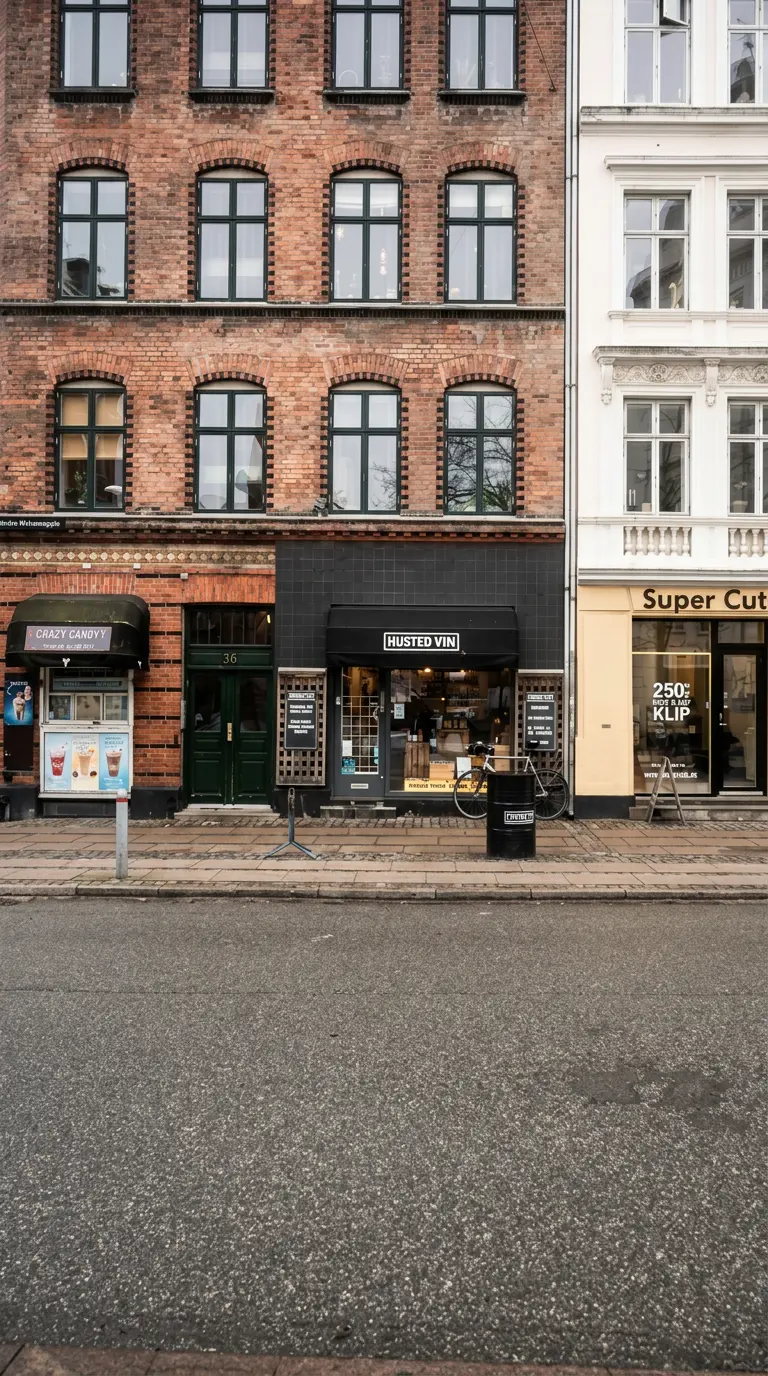 En tom gade med lagerbygninger og vinduer. En af bygningerne har en skilt med "Husted Vin" og en skulptur af en kvinde med is.