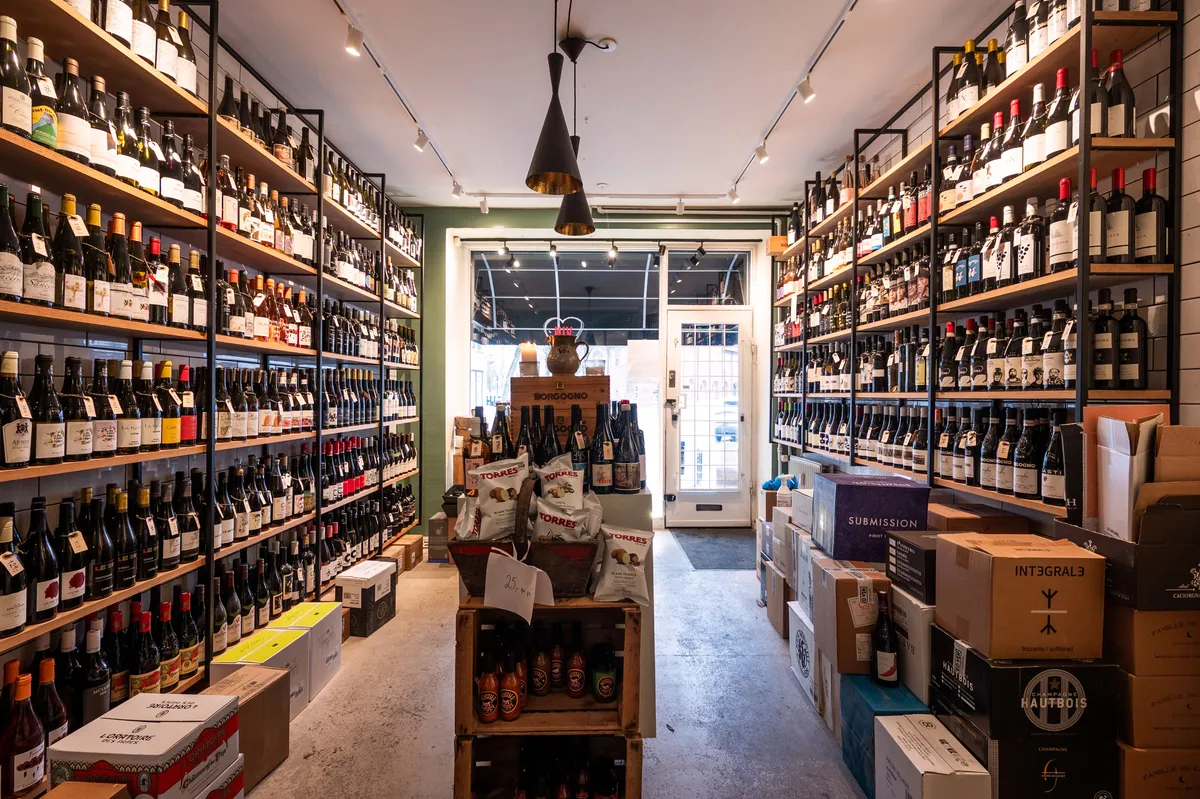 Vinbutik med udsnit af røde og hvide vinflasker og sager på hylder og gulvet og kasser af drikkevarer og snacks