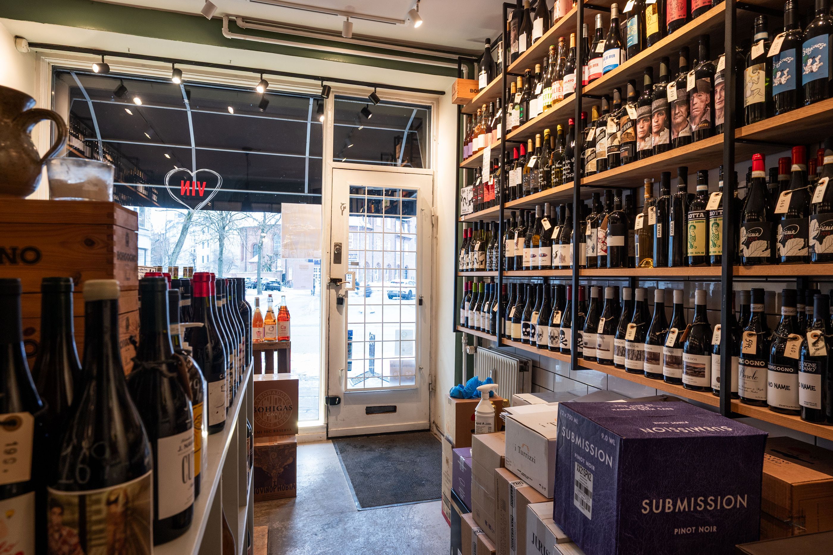 Butik med flere rækker vinflasker og etiketter samt udstilling af krukker og etiketter med indskriften Submission og Nossimios