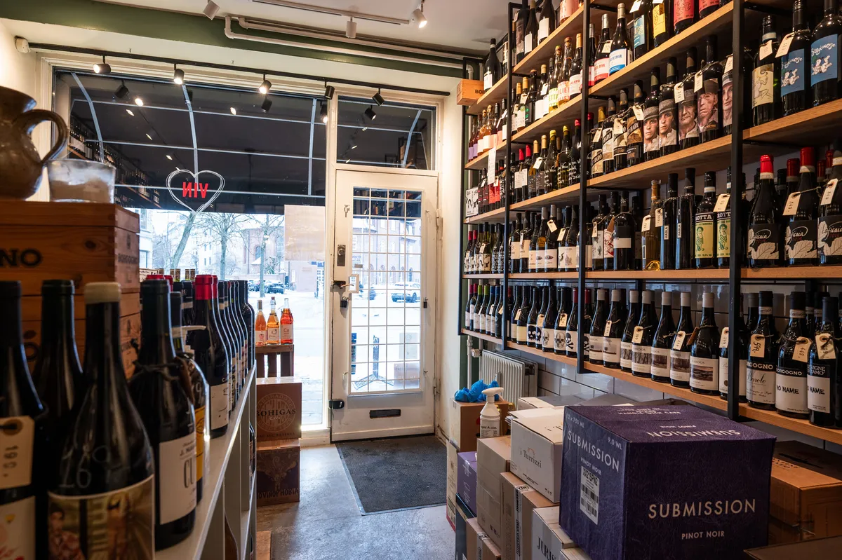 Butik med flere rækker vinflasker og etiketter samt udstilling af krukker og etiketter med indskriften Submission og Nossimios