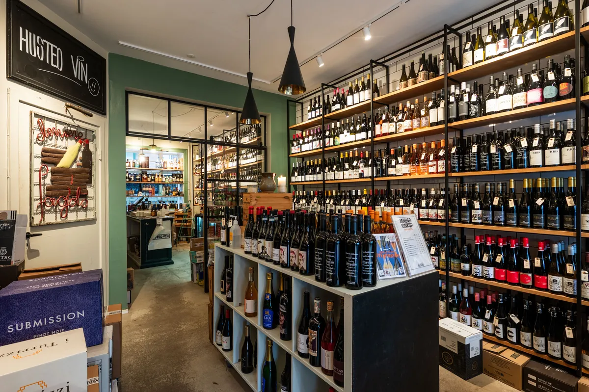 Butik med skovlformet lysekroner og hylder fyldt med vinflasker og etiketter. Der er et skilt med teksten Husted Vin på væggen og flere vinflasker og bokse på gulvet.