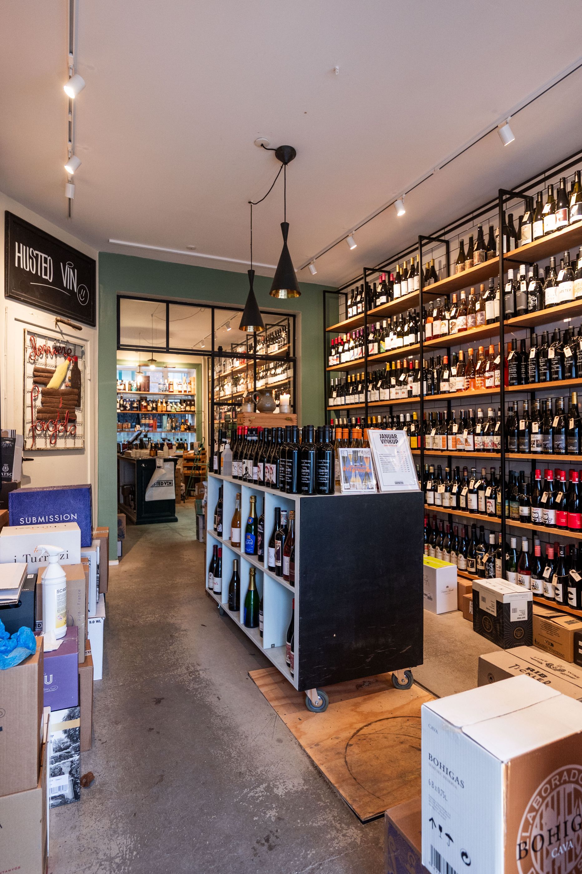 En vinbutik med store hylder med vinflasker og etiketter, lysekroner og sælger i bunden. Flaske vin på hjul og et sæt etiketter på bordet.
