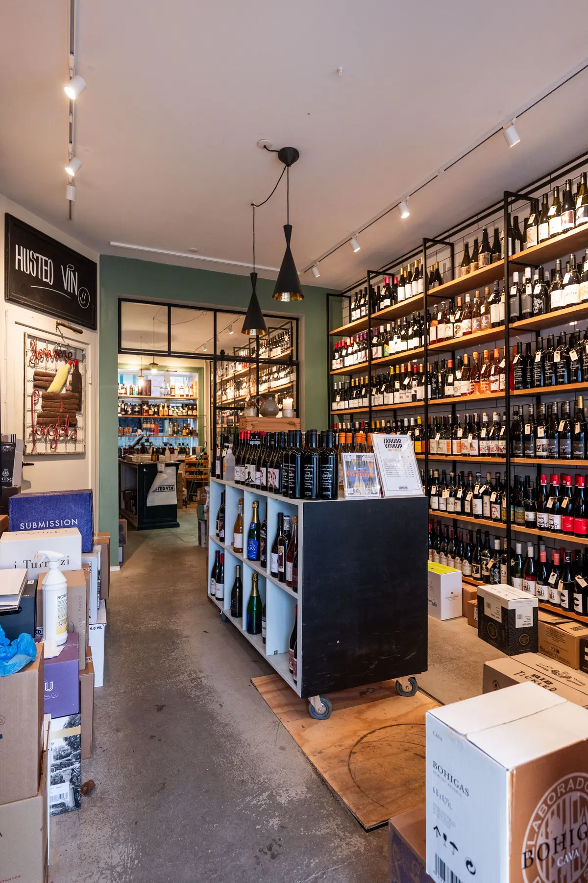 En vinbutik med store hylder med vinflasker og etiketter, lysekroner og sælger i bunden. Flaske vin på hjul og et sæt etiketter på bordet.