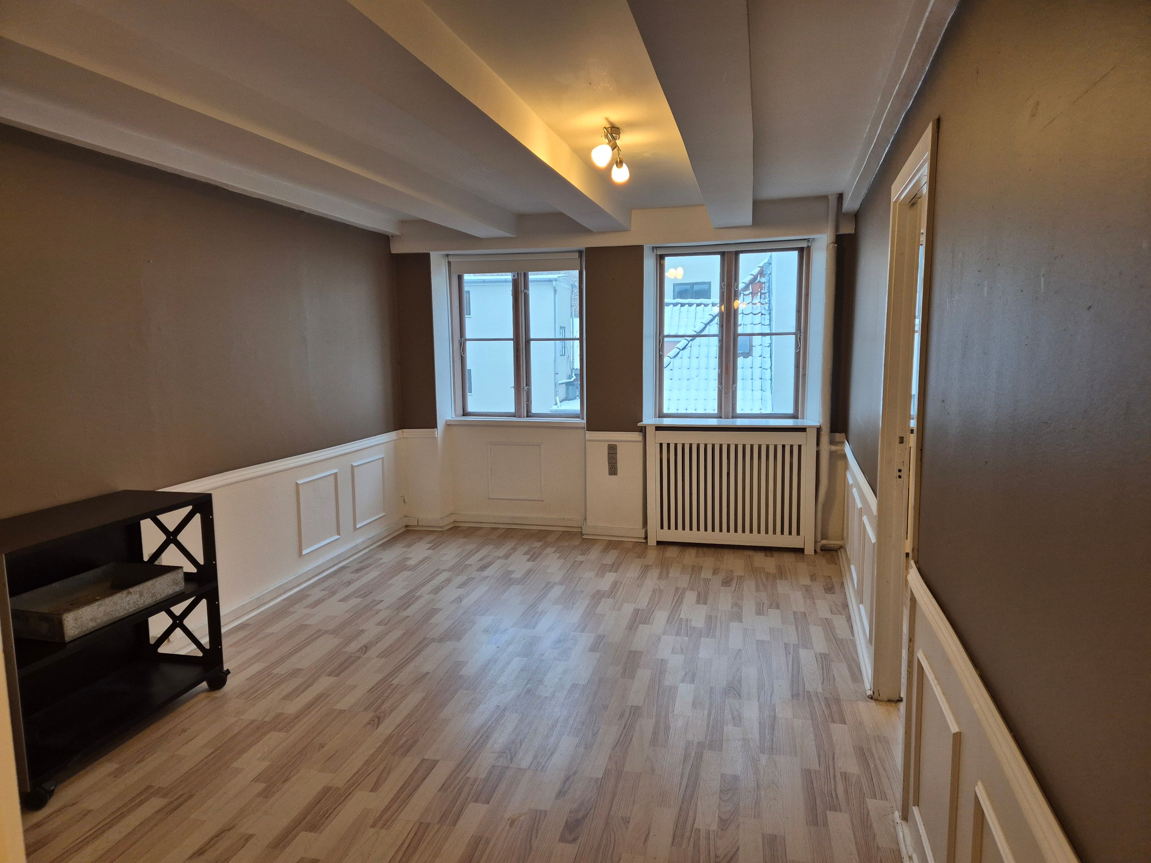 Kontor med højt til loftet, lyse gulve, to vinduer og radiatorer. En sort hylde med bokse og to lysepunkter på loftet