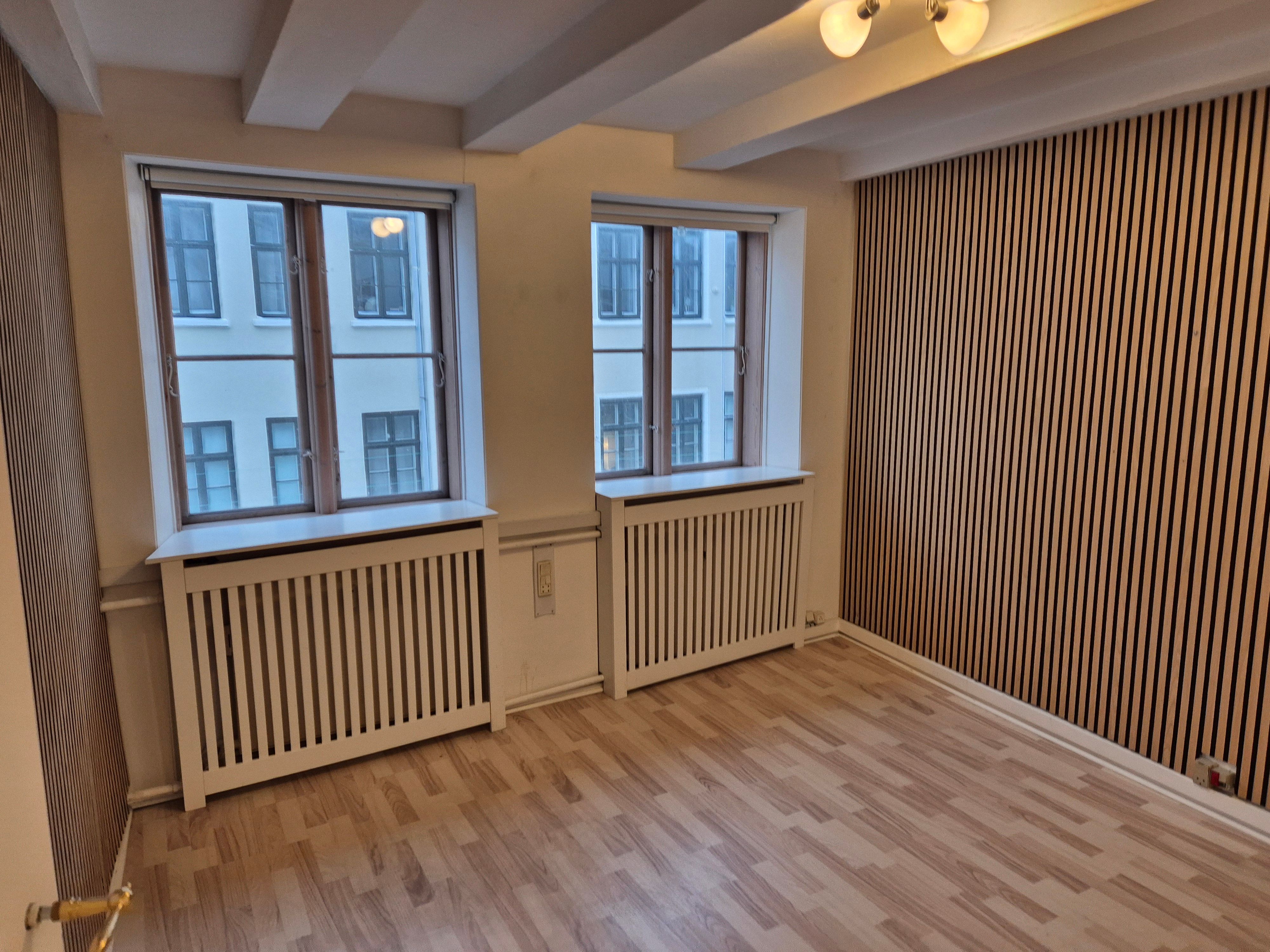 Lyst erhvervslokale med to vinduer og radiatorer på hver side. Træpaneler og lysekroner på loftet. Gulvbelægning i træ og strømstik på væggen.