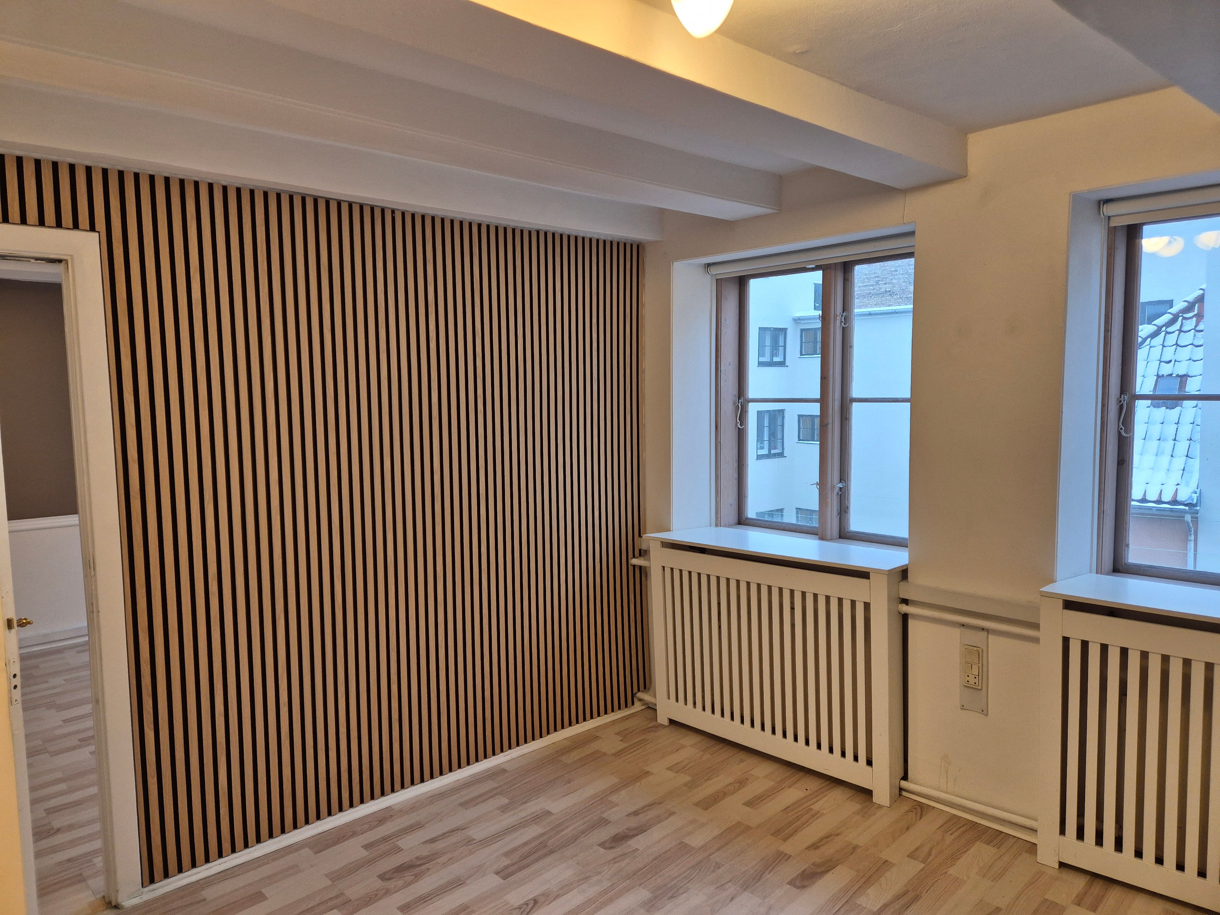 Et tomt kontor med to vinduer, radiatorer, højt loft, lysegule vægge og træpaneler på den ene væg