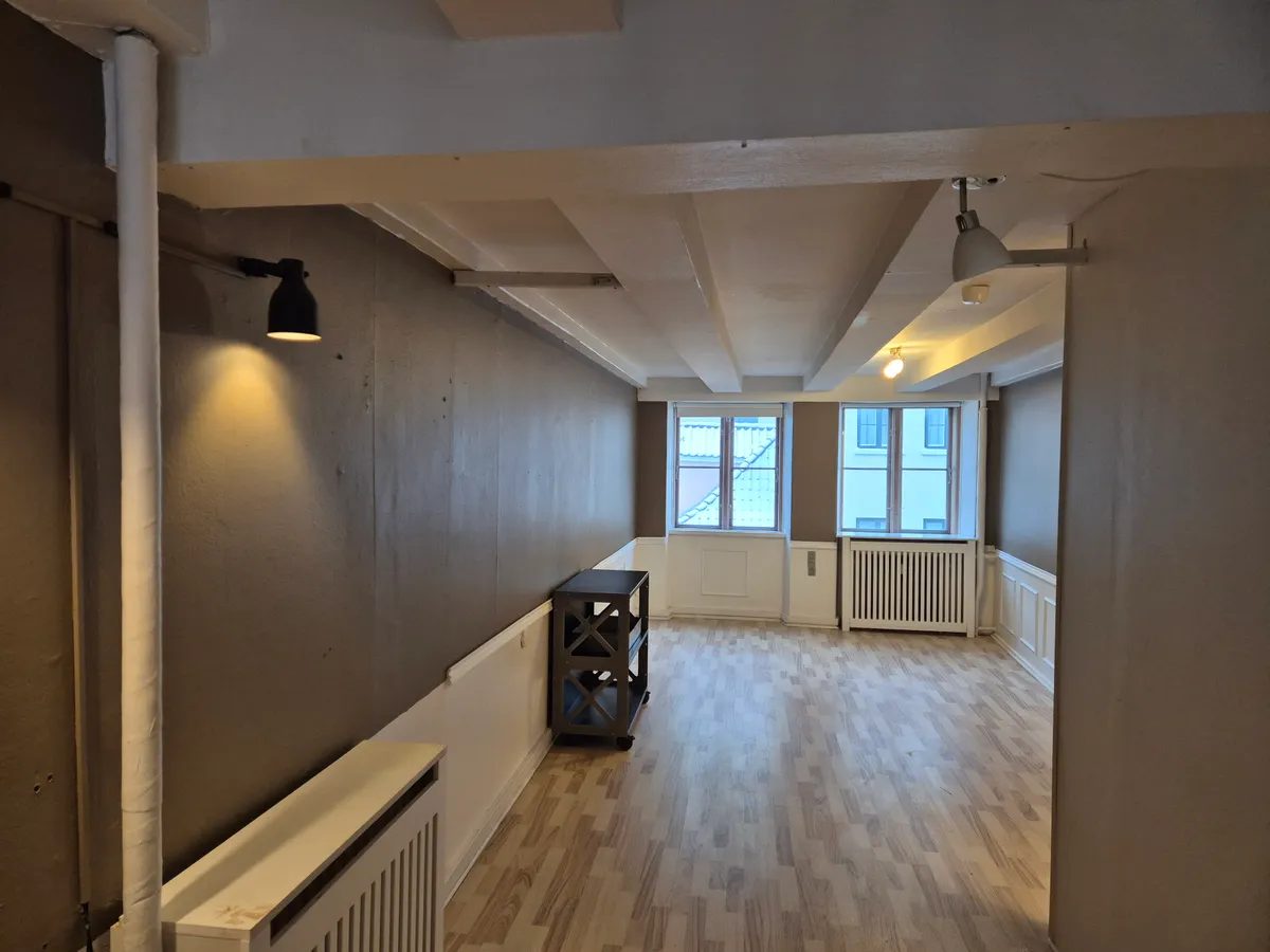 Kontor med lyst rum, højloft, trægulv og hvide vægge. Vægmontrerede lamper og radiatorer. En lille sort stol og to vinduer med udsigt til gårdhave.