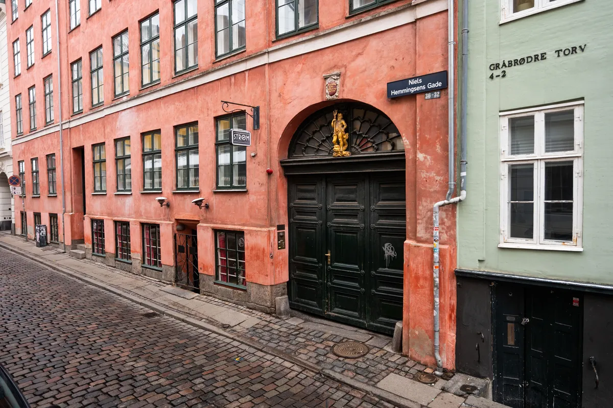 Værksted i Niels Hemmingsens Gade i København. Bygningen med røde vægge og store vinduer har lysegrå døre og en statue oven på porten. Der er skrifttægter og røre på væggene