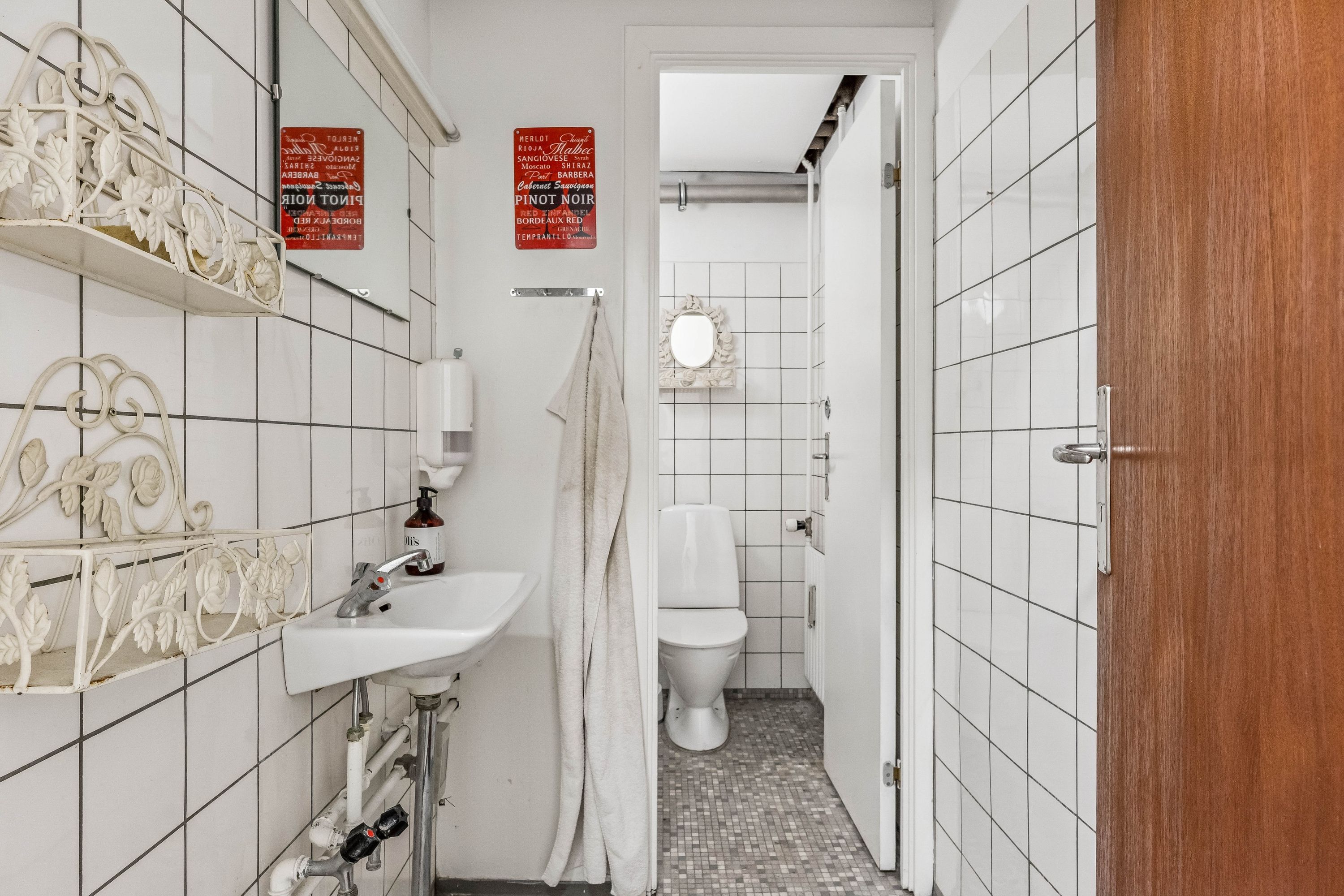 En lille og hvidklædt badeværelse med håndklæde, håndvask og toilet samt udsmykning i form af røde skilte med alkoholnavne