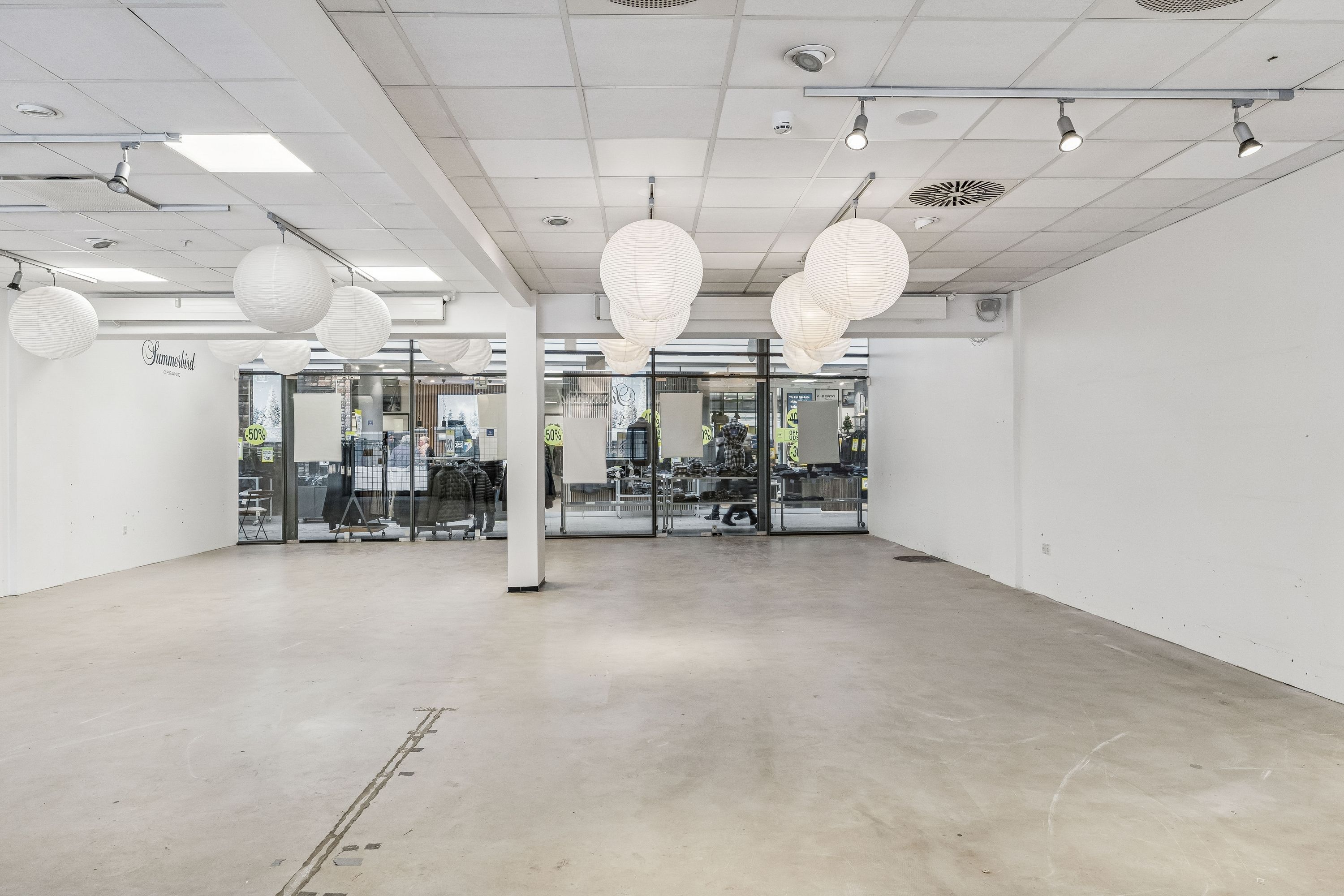 Rummelig butikslokal til leje med lyse gulve og hvide vægge, hængende paplampe og glasdøre til lagerrum. Der er en udsigt til en butik med klæderne i udsalgsvinduet.