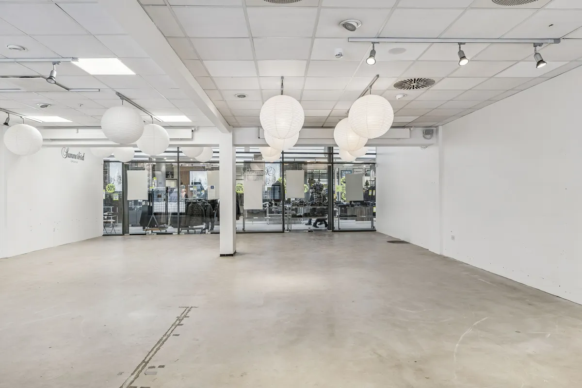 Rummelig butikslokal til leje med lyse gulve og hvide vægge, hængende paplampe og glasdøre til lagerrum. Der er en udsigt til en butik med klæderne i udsalgsvinduet.