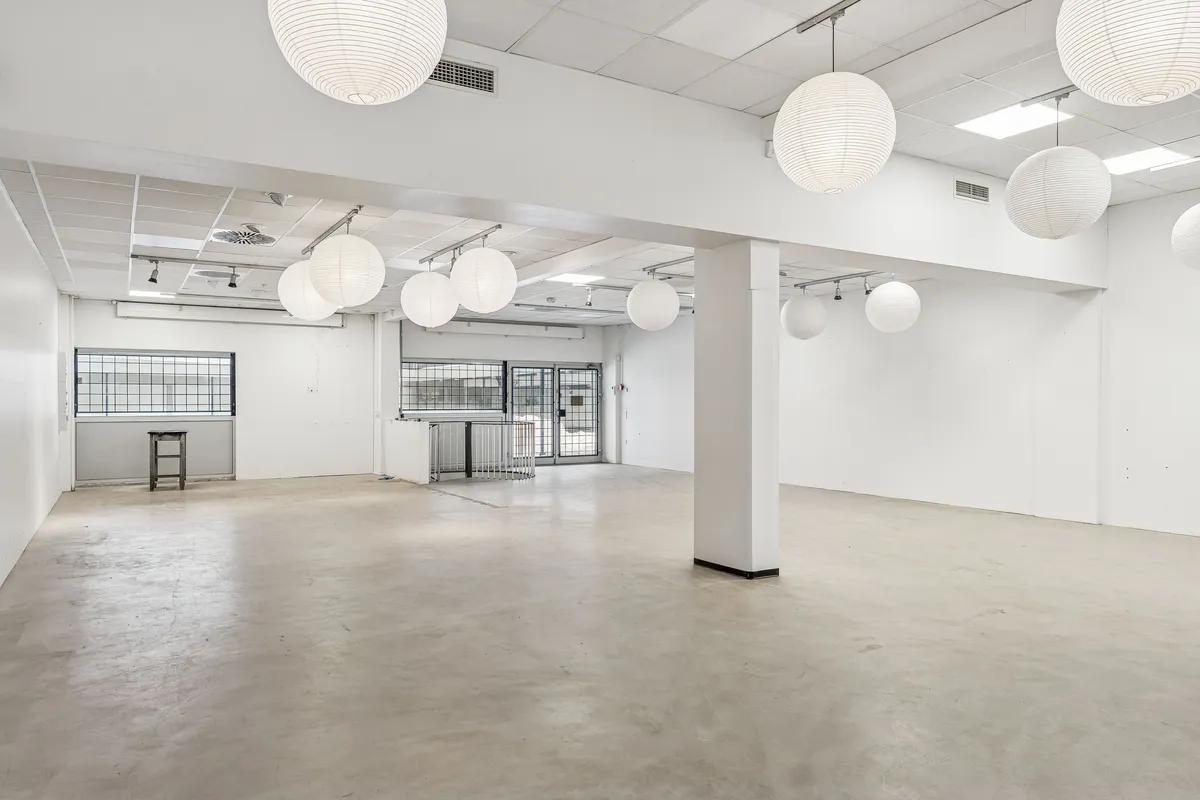 Store og veldisponerede butikslokaler med hvide vægge og lysekroner, der giver god belysning. Lukket skur og glasruder med stangsikring. Betongulv og højloftet design med lysekroner i loftet.