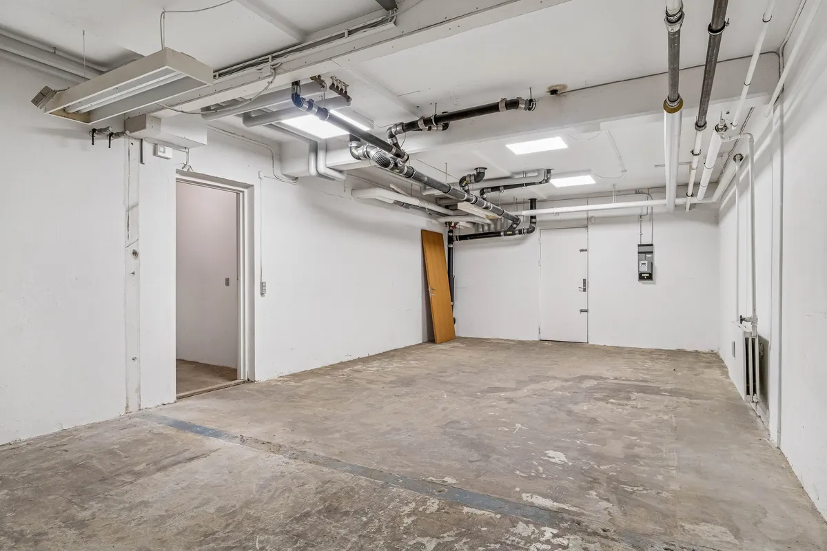 Store butikslokaler med kælder og flere indgange, slanke lysekroner, rørsystem og væghylder, en åben dør og en lukket dør, hvide vægge og gulv og lysepunkter