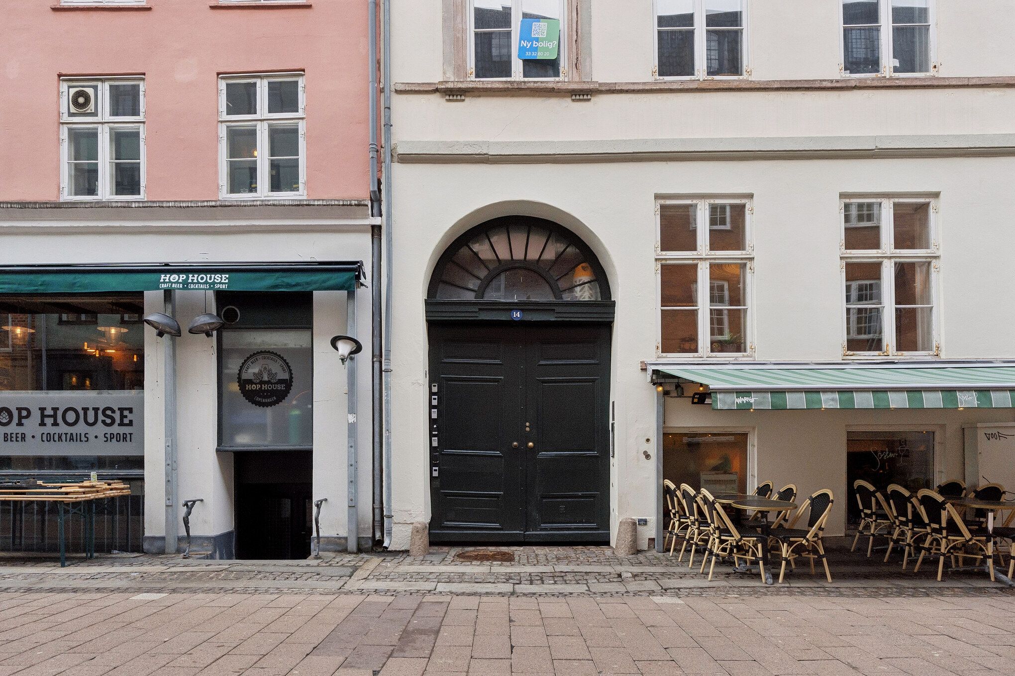 Bygninger med vinduede facader og indgange i Kompagnistræde, et kvarter med cafeer og småforretninger, tæt på Gammel Strand metrostation. Højre bygning har grøn-hvide aubussoner og stolpeborde med stole.