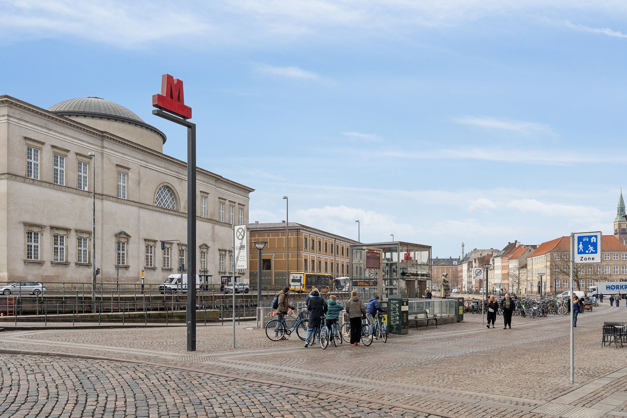 En stor bygning med røde bogstaver og et kuppelformet tag. Folk cykler og går rundt på Strædet. Der er også parkerede cykler og en stor bus i baggrunden.