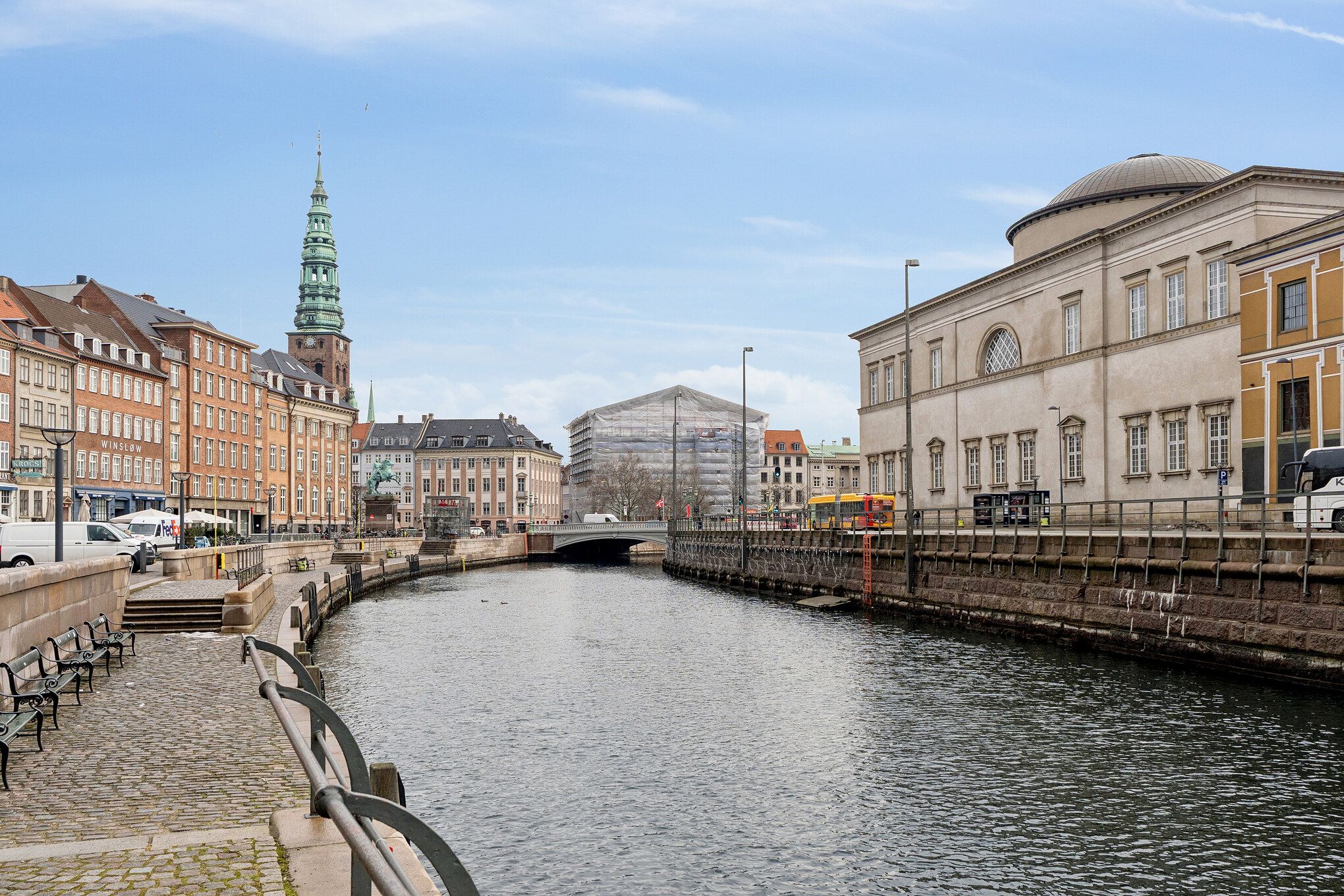 Kontor i det charmerende Strædet med udsigt over vandløb og bygninger, busser og bænke langs vandet, og slanke maste og gamle røde bygninger i baggrunden