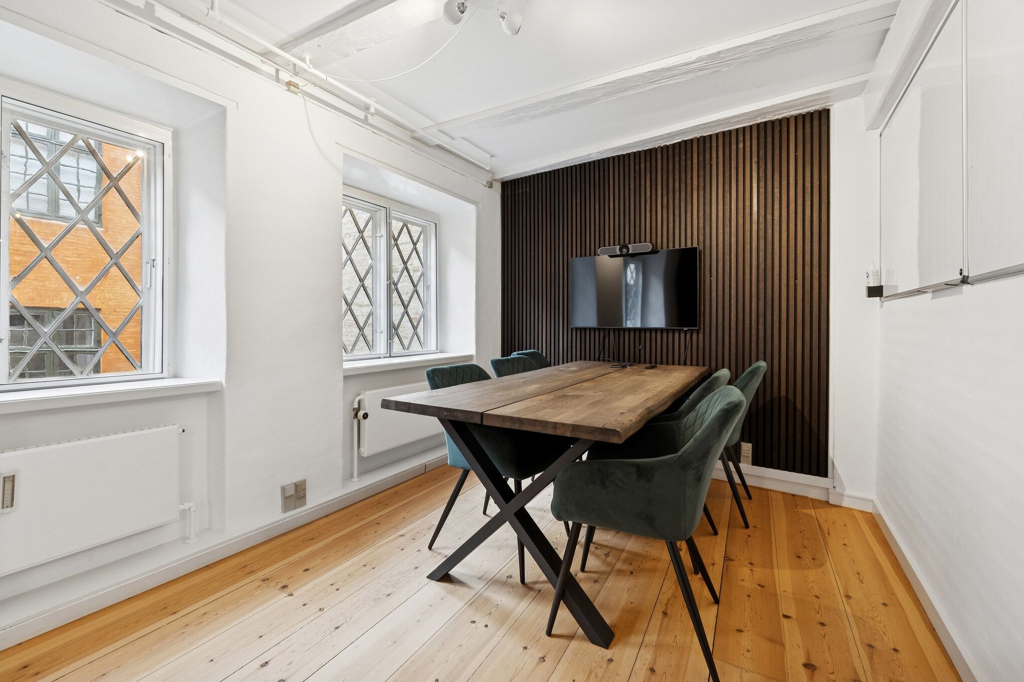 Mødelokalet med lyse trægulve og lyse vægge har lysegrå stole og et langt bord med krydsben. Der er to vinduer med krydsrute og en tv-skærm på væggen. Loftet er hvidt og der er en radiator under vindueskarme.