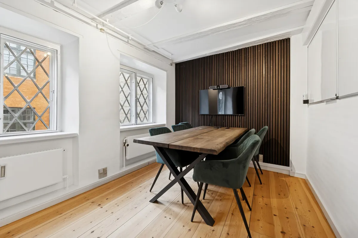 Mødelokalet med lyse trægulve og lyse vægge har lysegrå stole og et langt bord med krydsben. Der er to vinduer med krydsrute og en tv-skærm på væggen. Loftet er hvidt og der er en radiator under vindueskarme.