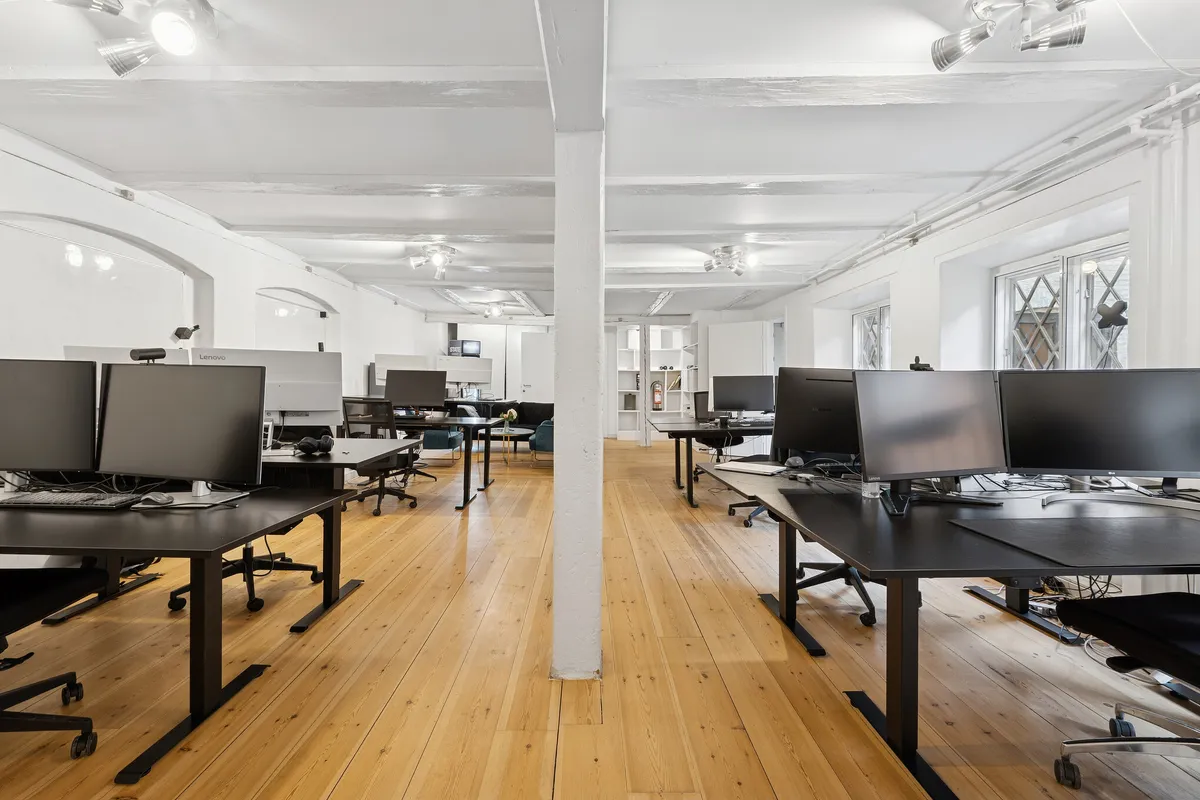 Moderne kontor med trægulve, hvide vægge og møbler, flere skriveborde med monitorer, stolpe og lysekroner i loftet, opbevaringsbokse og glasvinduer i baggrunden