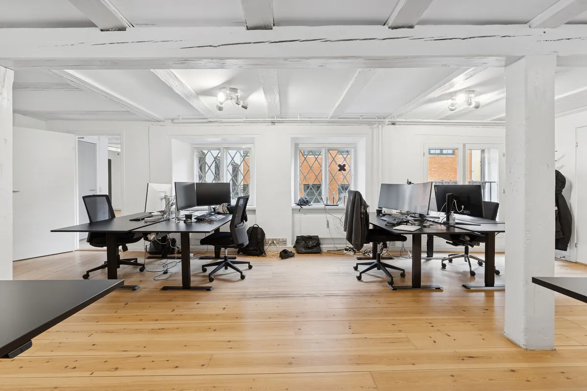 Kontorrum med lyse lofter, moderne møbler, computere og skriveborde, alle fordelt i et stilrent og renset miljø med trægulve