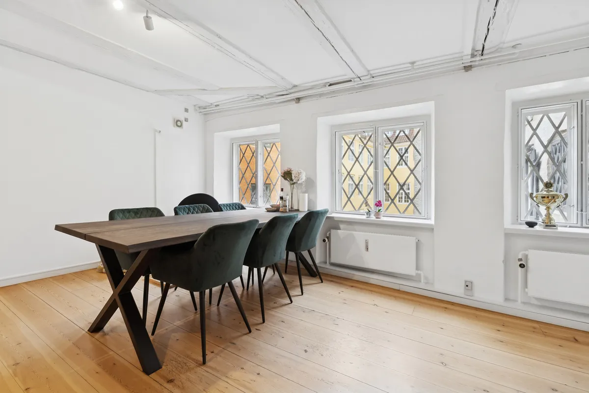 Kontorlokaler med lyse trægulve, hvide vægge og store vinduer med udsigt til en bygning med historiske mønstre. En lang træbord med grønne stole og en vase med blomster på vindueskarme