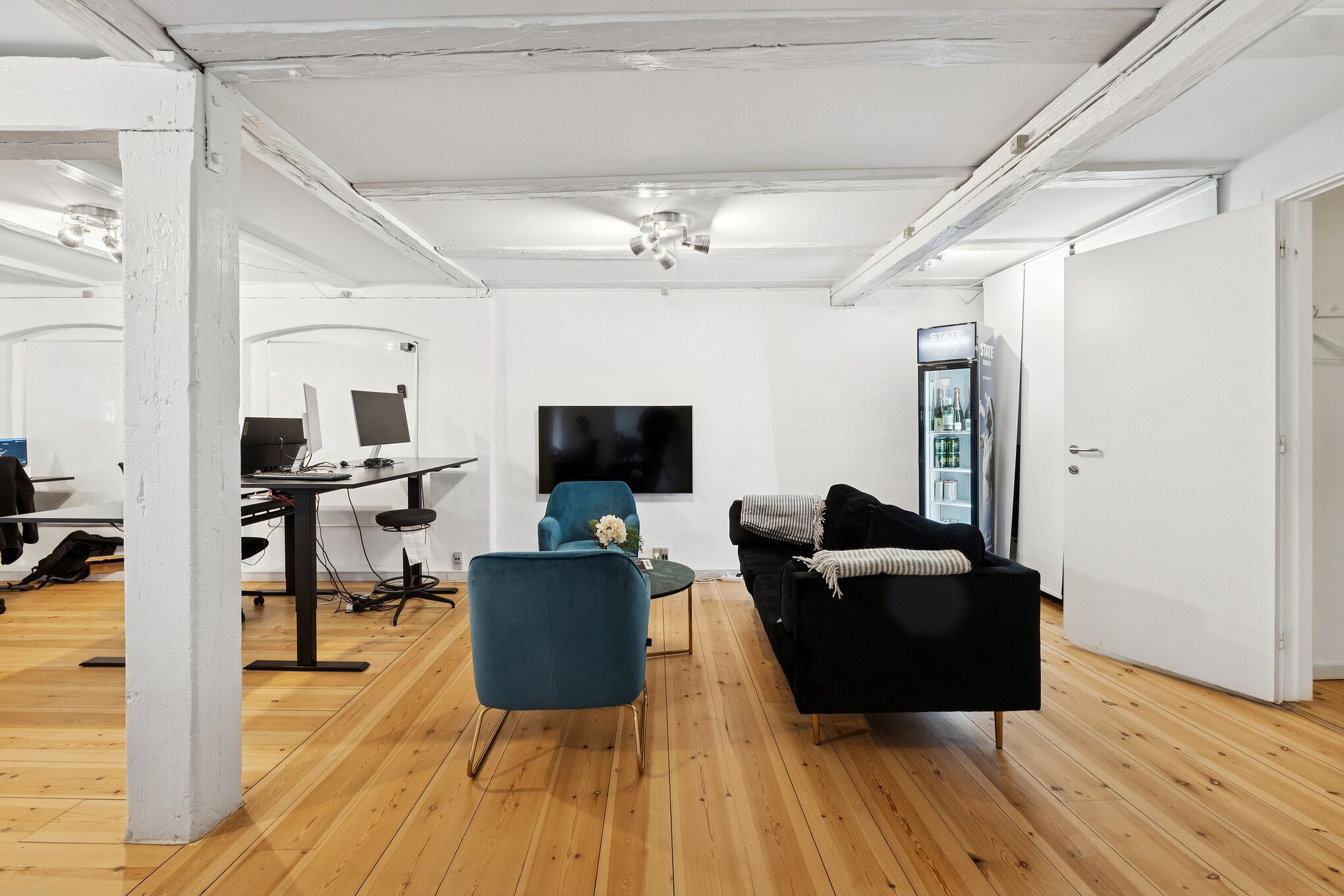 Kontor med højloft, sofa, bord med skærme og højtalere, køleskab og højttalere på loftet, gulv med lyse træplanker