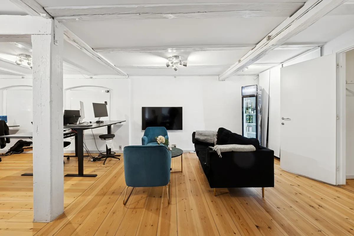 Kontor med højloft, sofa, bord med skærme og højtalere, køleskab og højttalere på loftet, gulv med lyse træplanker