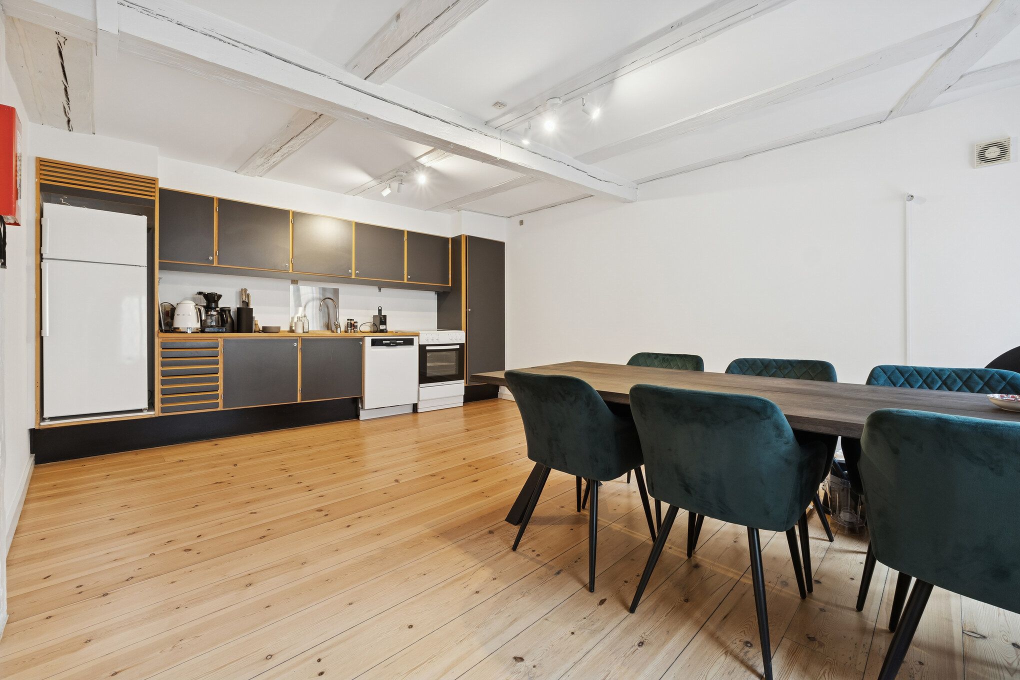 Moderne køkken med hvide vægge og lysegrå loftsbjælker, hvidt køleskab og ovn, lysegrå krydsfiner og skarpe, grønne stolene rundt om et langt træbord