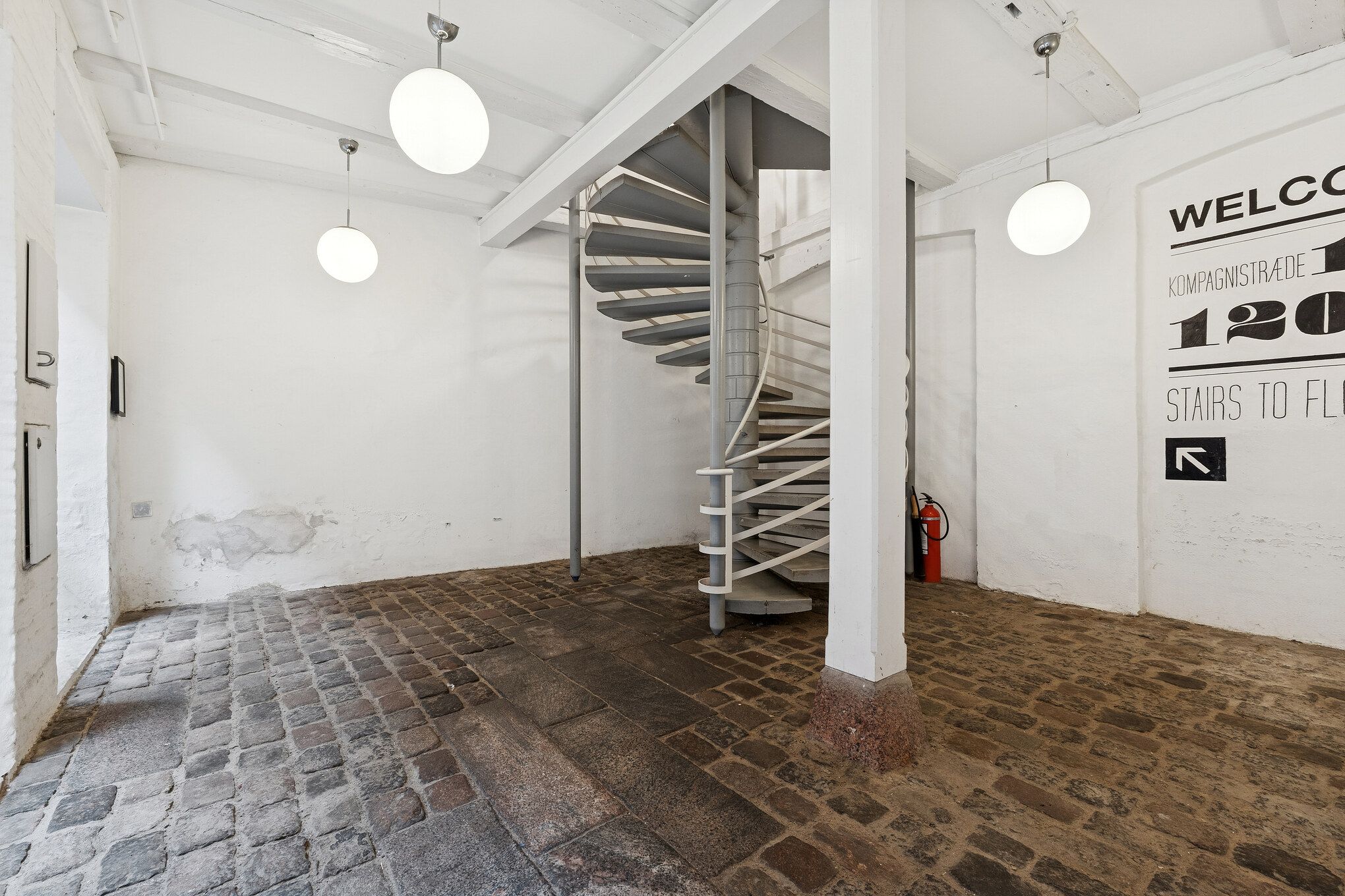 Kontorrum med åben indgang, spiraltrappe, gulv af sten og tre hængende rundt lys. Væg med velkomstskilt og brandudstyret.