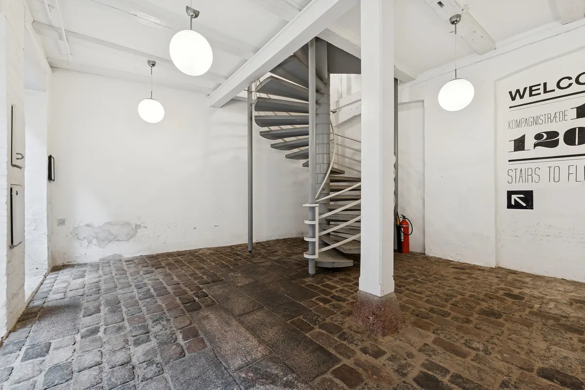Kontorrum med åben indgang, spiraltrappe, gulv af sten og tre hængende rundt lys. Væg med velkomstskilt og brandudstyret.
