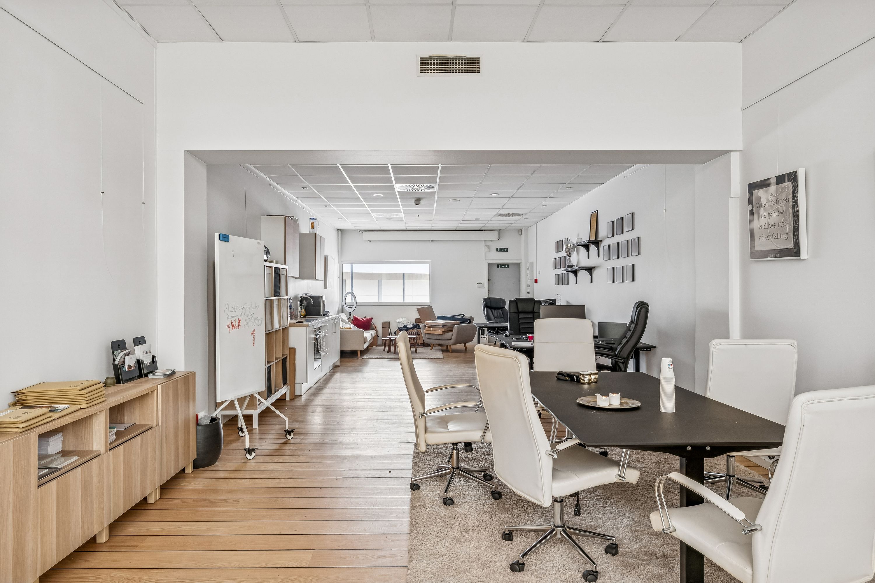 Hvidt kontorlokale med lysegul trægulv og hvide stole med hjul. Kontorbord, skrivebord, skrivemaskine og fotoramme på væggen. Forskellige møbler og stole placeret i lokalet.