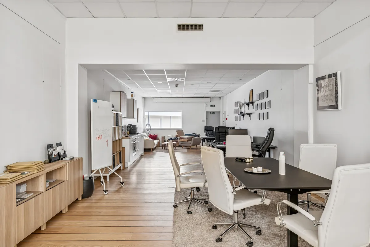 Hvidt kontorlokale med lysegul trægulv og hvide stole med hjul. Kontorbord, skrivebord, skrivemaskine og fotoramme på væggen. Forskellige møbler og stole placeret i lokalet.