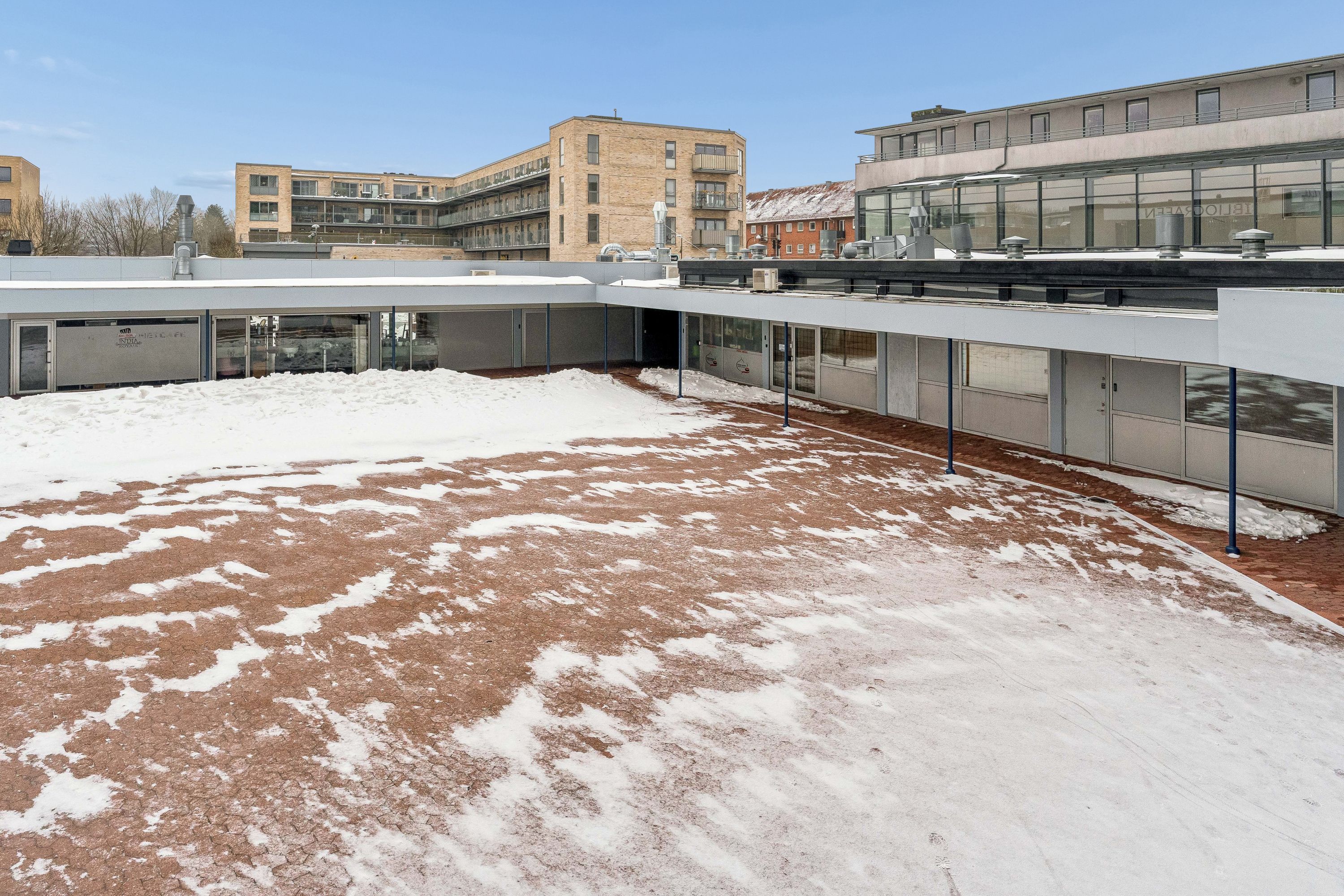 Et lejlighedskompleks med en terrasse og en gårdhave dækket i sne og rødt gulv, med moderne bygninger og glasvinduer i baggrunden, og sne på taget