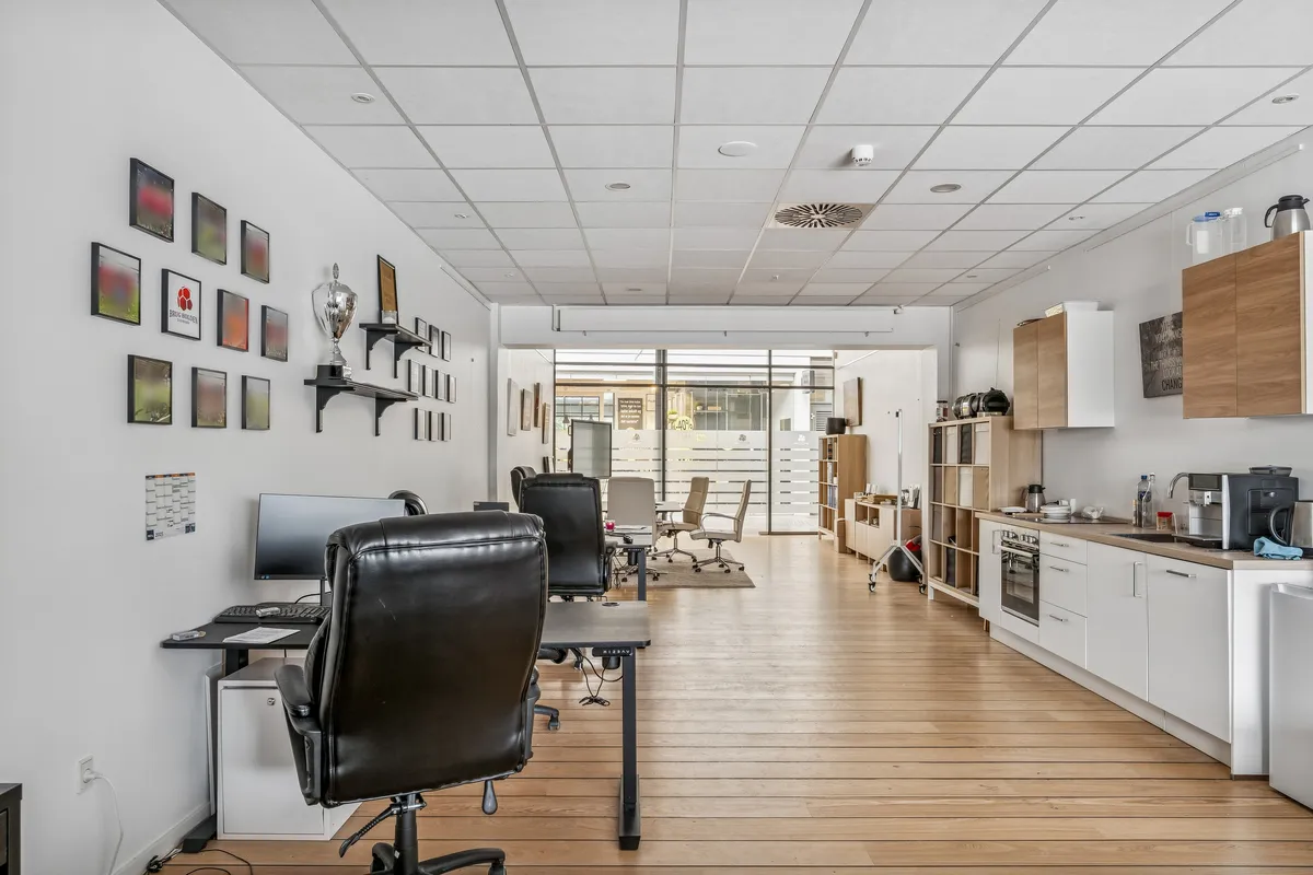 Kontor med lyse trægulve, moderne kontormøbler og teknologi, såsom computere og skærme. Bagved er der et lille køkken med en ovn og et køleskab.