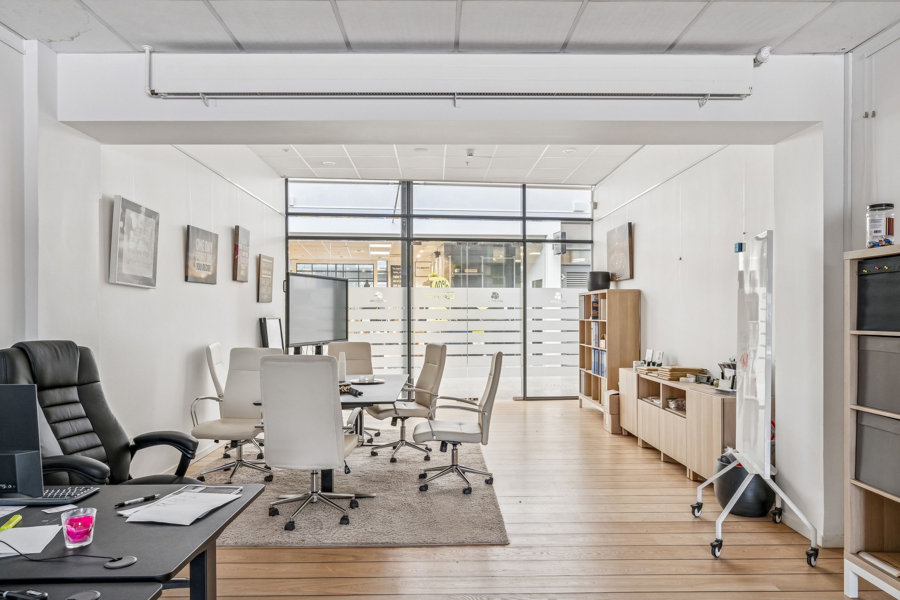 Foto af et moderne mødelokale med trægulv, højt loft, lyseruder og minimalistisk indretning. Møbler inkluderer konferencebord, lyse stole og en stor skærm.