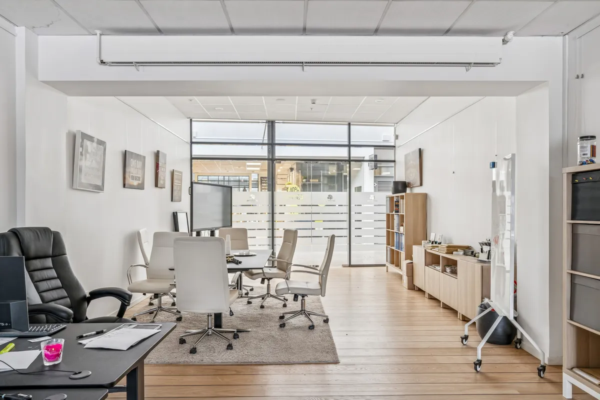 Foto af et moderne mødelokale med trægulv, højt loft, lyseruder og minimalistisk indretning. Møbler inkluderer konferencebord, lyse stole og en stor skærm.