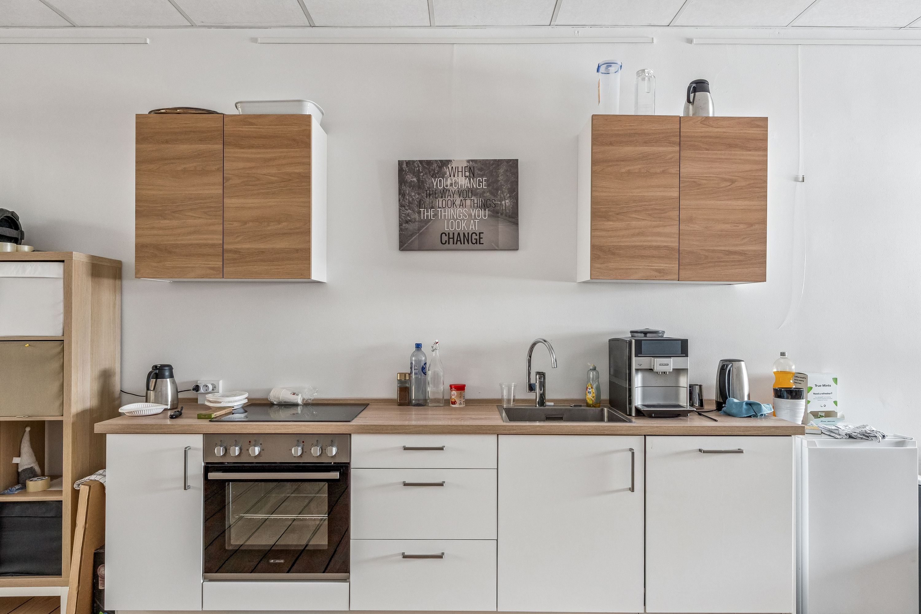 Regleret køkken med lyse vægge, træskabe og moderne indretning. Et svart kaffemaskine, en ovn og en vandhane med lyserødt vand er synlige.