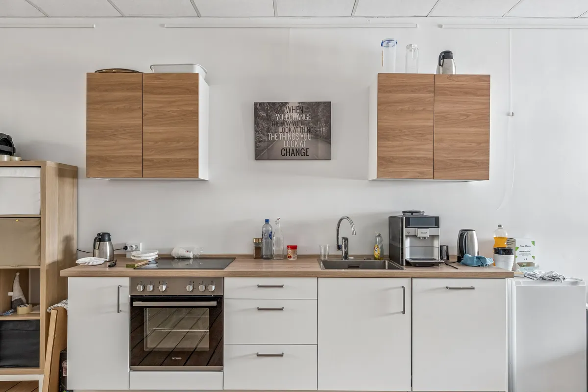 Regleret køkken med lyse vægge, træskabe og moderne indretning. Et svart kaffemaskine, en ovn og en vandhane med lyserødt vand er synlige.