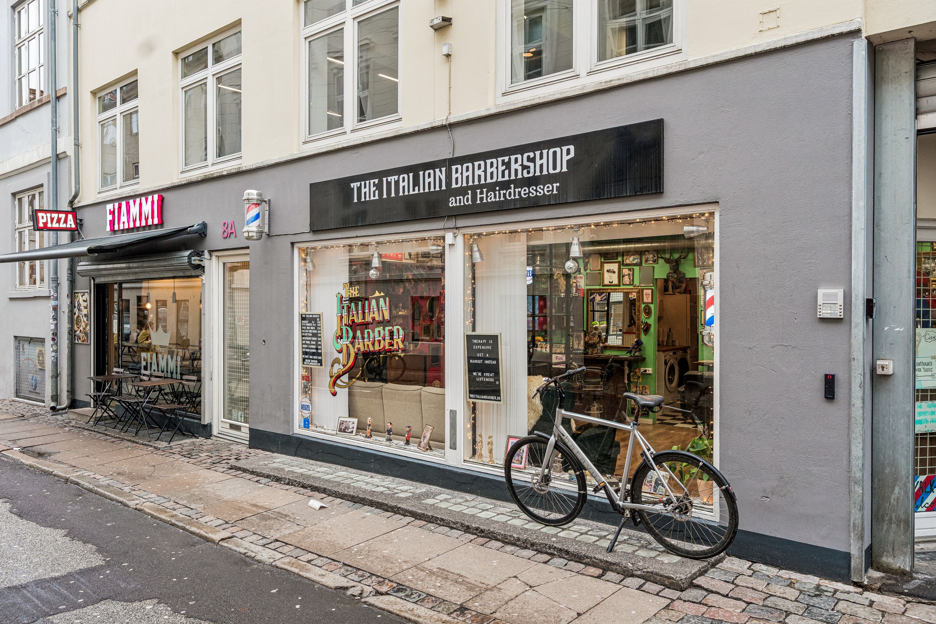 Cykel parkeret foran The Italian Barbershop og Hairdresser på Knabrostræde i København, med skrifttavle og udstillingsvindue, pizzarestaurant med rødt skilt og spisestedets bord og stole på gågaden