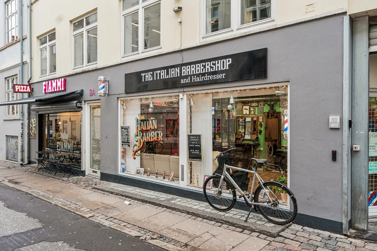 Cykel parkeret foran The Italian Barbershop og Hairdresser på Knabrostræde i København, med skrifttavle og udstillingsvindue, pizzarestaurant med rødt skilt og spisestedets bord og stole på gågaden