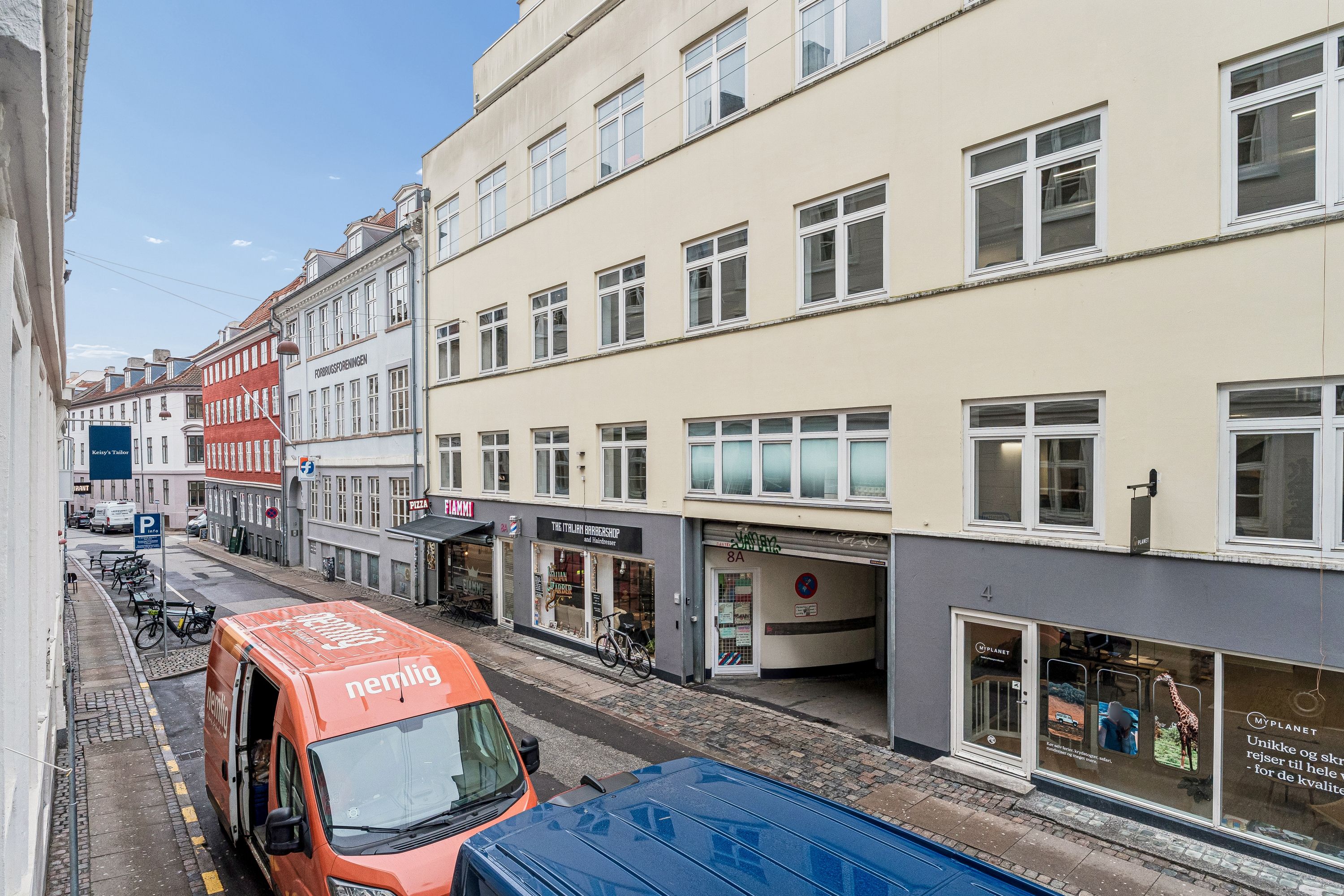 To orange og blå varevogns kører på Knabrostræde, og butikker med vinduer er på hver side af gaden. På hjørnet er en plakat med en giraffe.