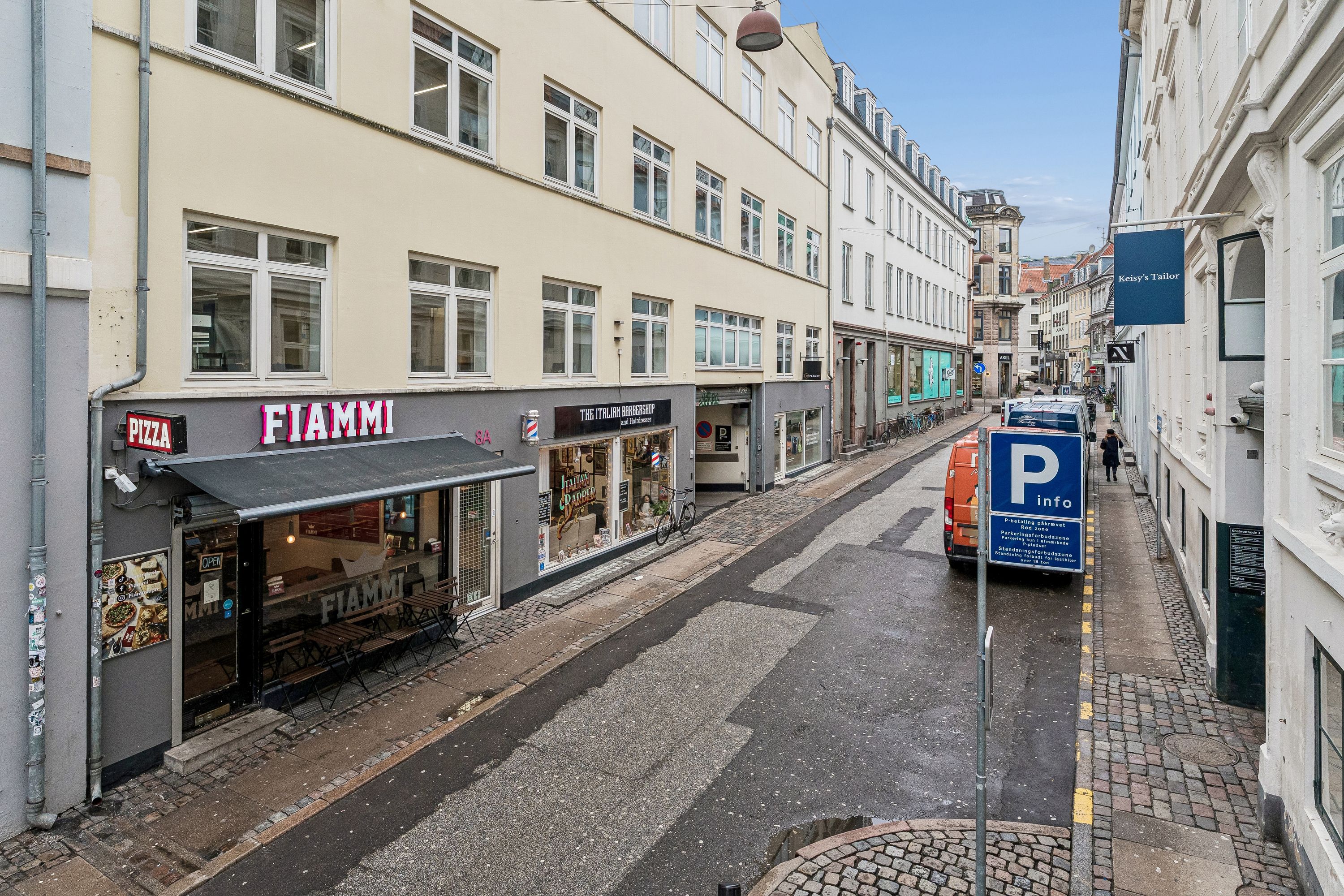 Københavnsk gade med forretninger og huse. Et parkeringsskilt står på siden af vejen. En bil parkeret på gaden. En person går langs fortovet. En fiammede pizza skilt og The Italian Barbershop butik.