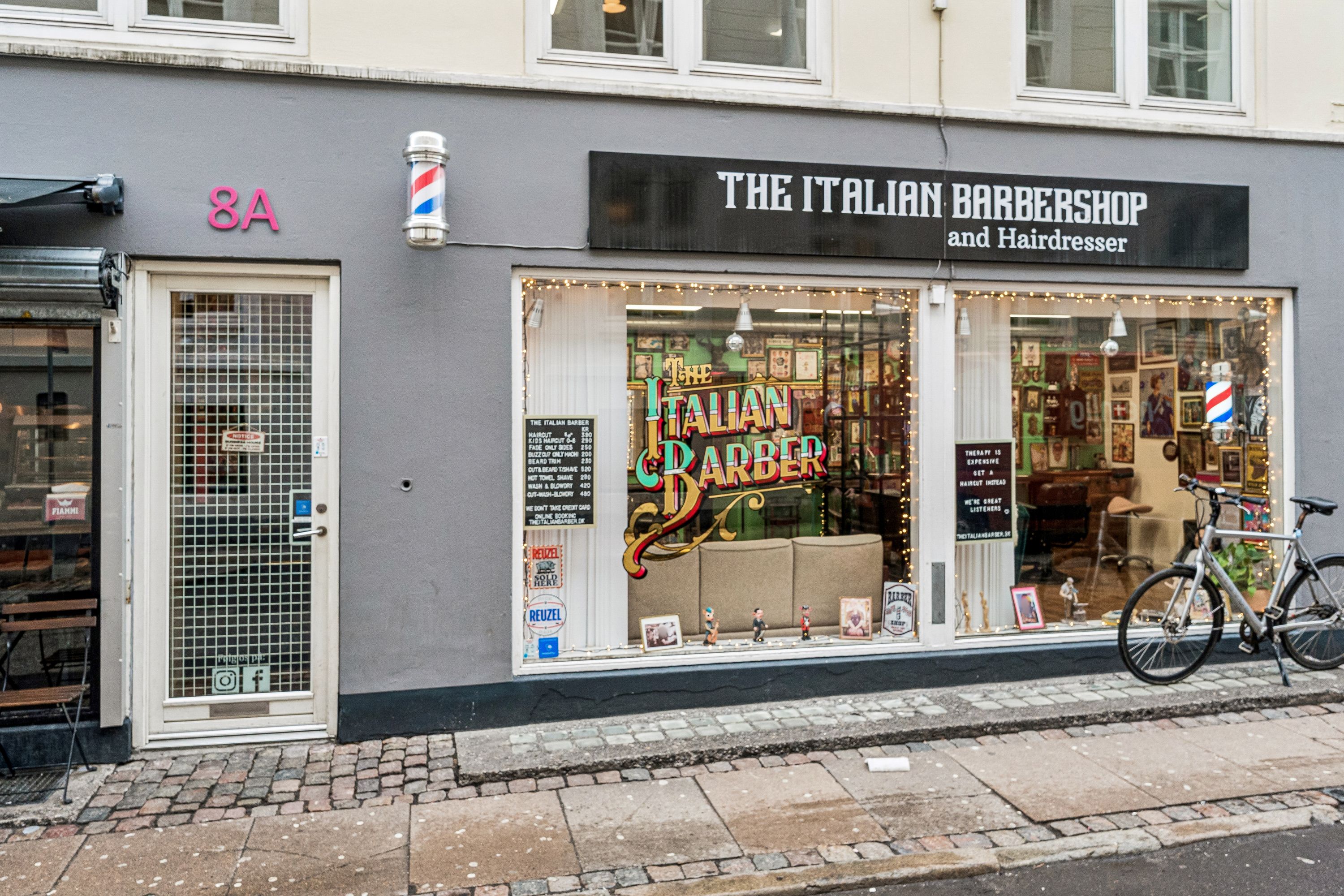The Italian Barbershop og frisørforretning i Knabrostræde 8A, København K. Med barberpole og briller i vinduet og en cykel parkeret udenfor.