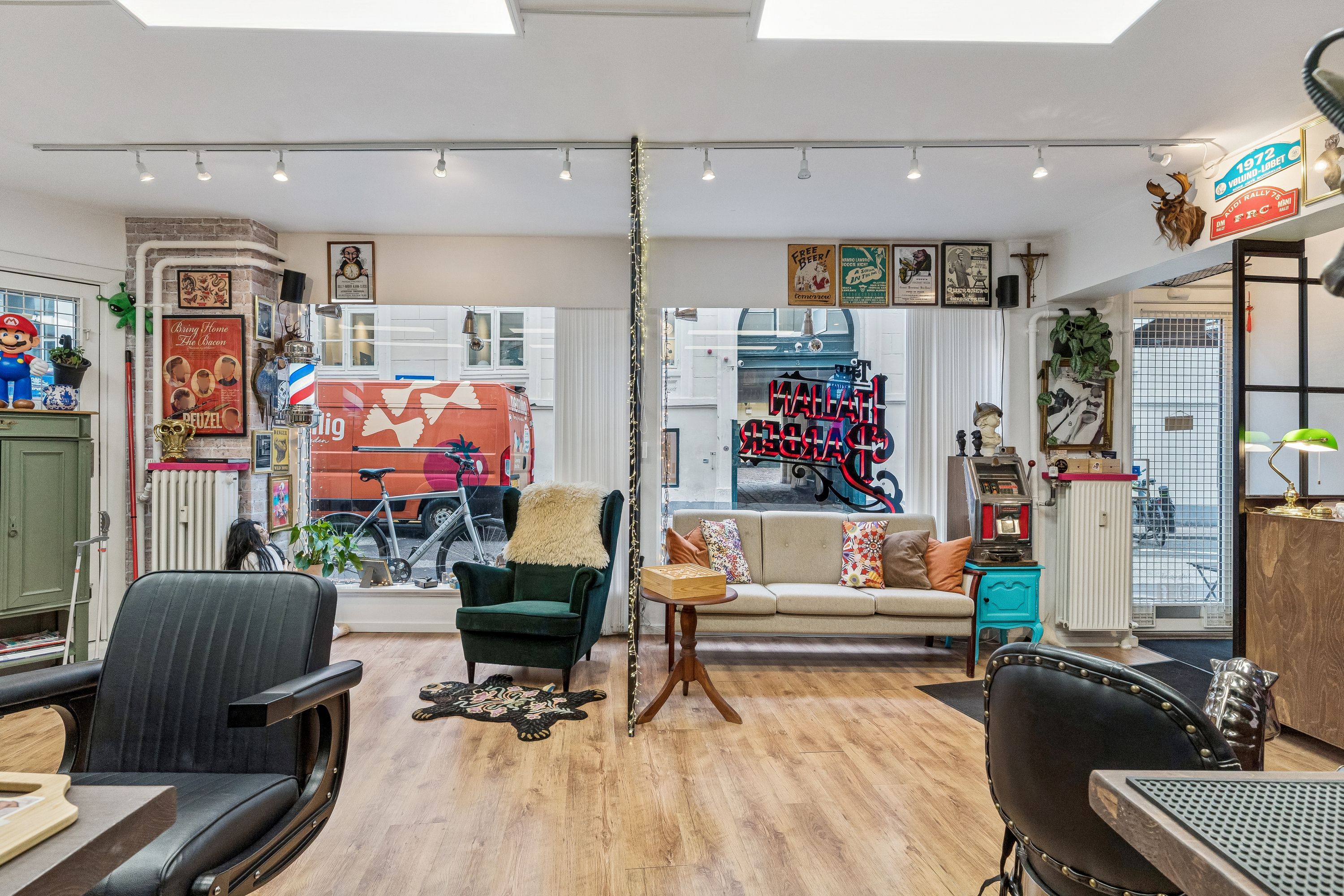 Butik med lyse trægulve, moderne møbler og vægmaleri, hvor der er en sofa og en barok stol med puder, og der er lysekroner og kunstværker på væggen