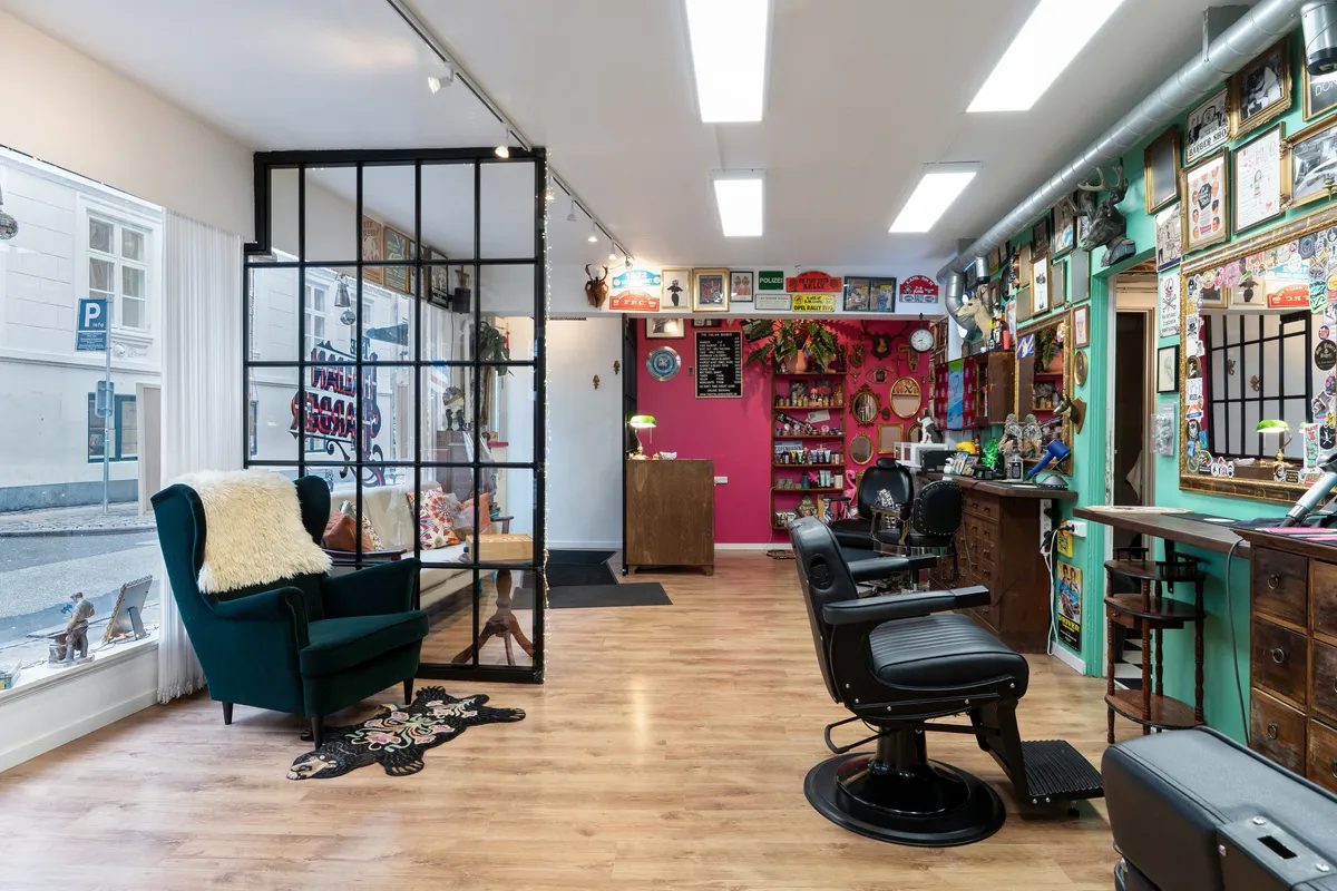 Barbersalon med barberstole, smykkeskabe og malerier. Lamper på loftet og spejl. Lyserød væg med et ur og plante.