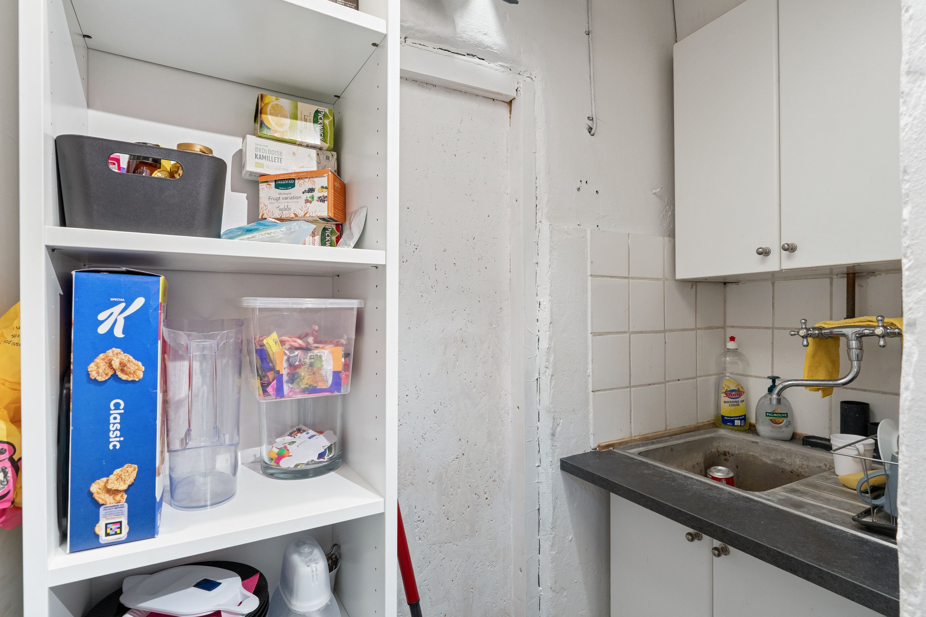 En køkken med en hvid hylde, krydderibokse og skåle, samt et rent vaskeblik med sæbe.