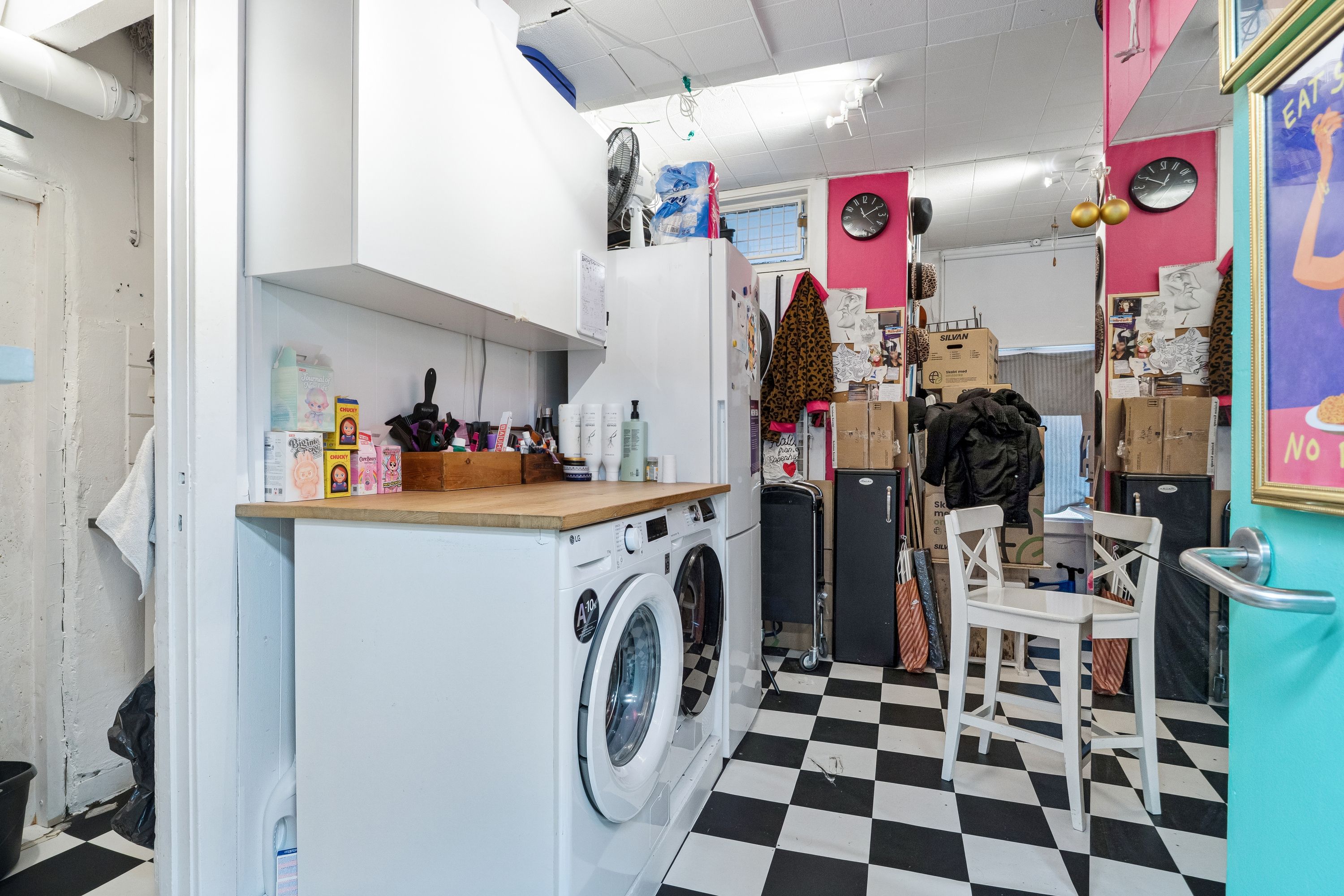 I stuen er der et hvidt vaskemaskine og tørretumbler samt en køleskab med kufferter og ting på gulvet og væggen med klokker og malerier