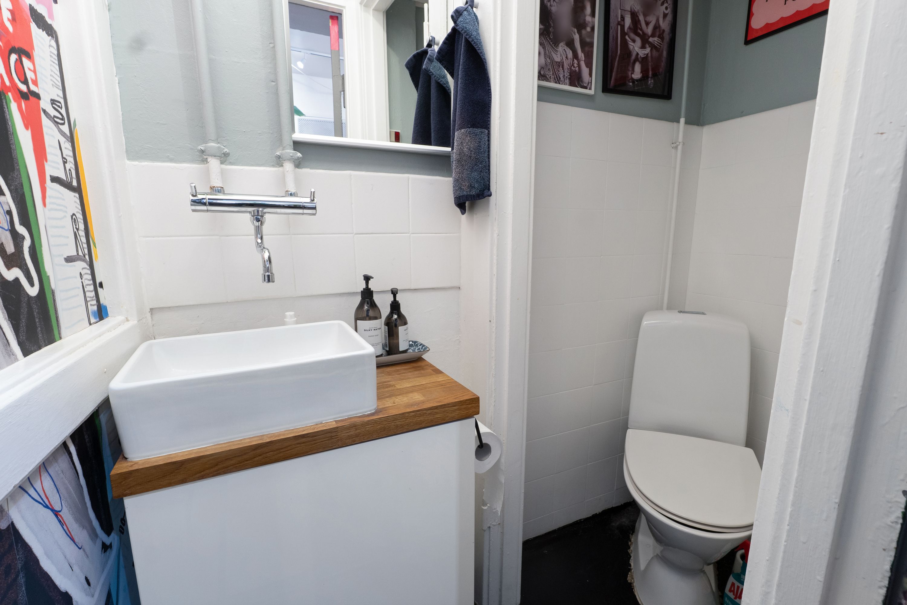 Et lille hvidt badeværelse med to toiletflasker på vaskentøj, hvidt toilet, toiletpapir og højt op til loftet. Hvidt hjørneskab med sink og vandhane samt to kunstværker på væggen