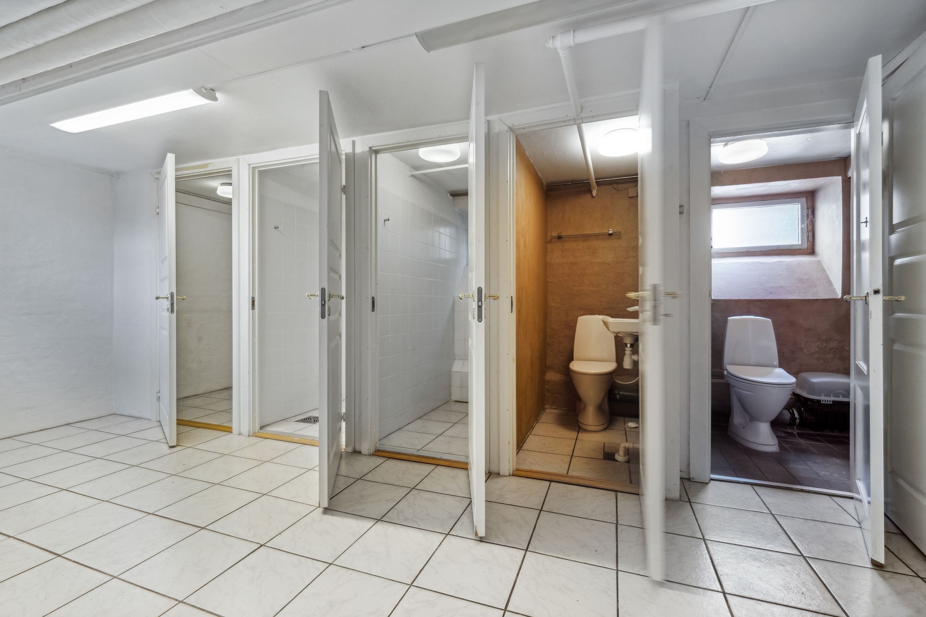 Et stort rum med hvide vægge, hvor der er to toiletter med hvide toilettæpper, og en hvid badeværelseskabinet med hængende lys